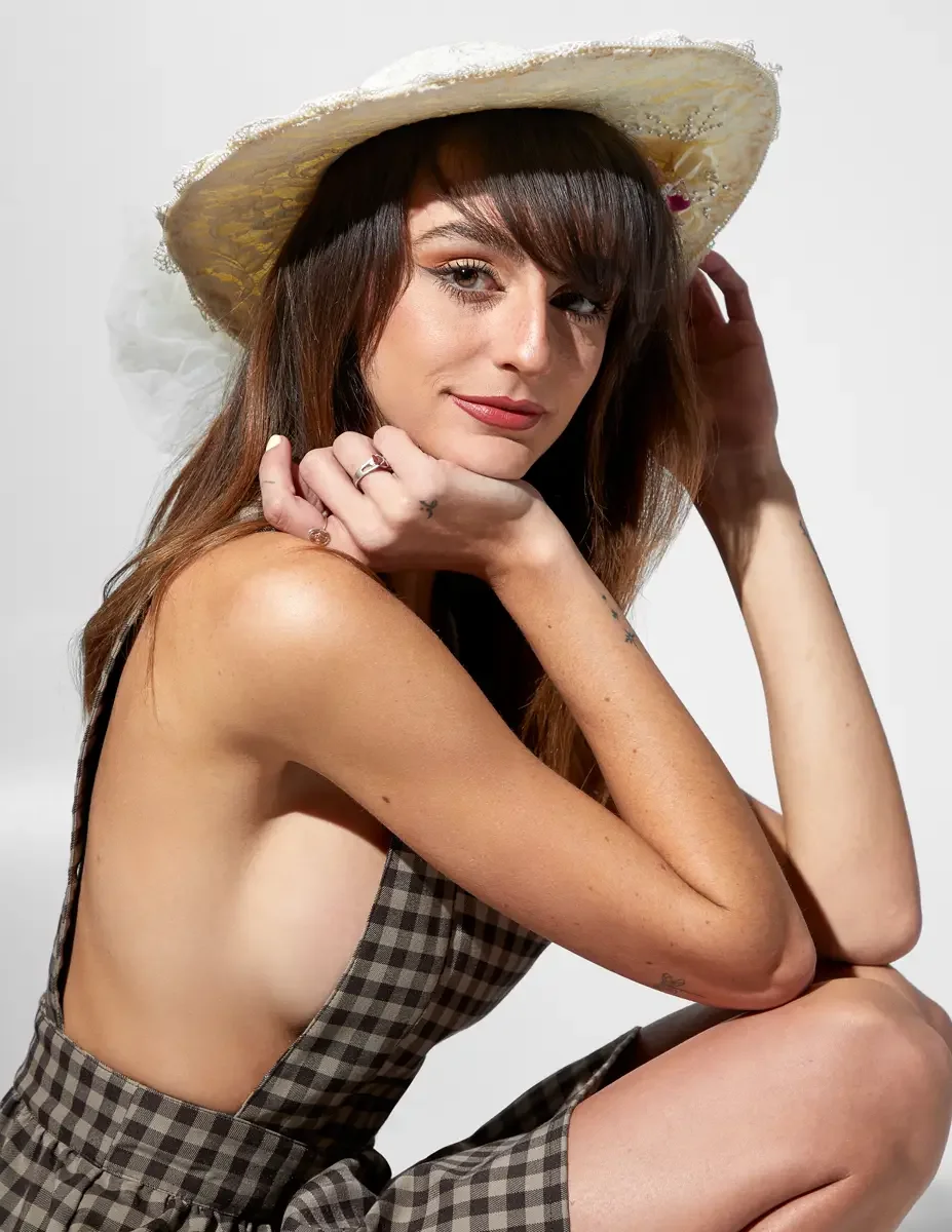 Macy, close-up portrait, seated, hands at chin, brown and black gingham tiered midi dress with open back, cream wide-brim straw hat with white tulle and floral trim, small tattoos on forearm and wrist visible on this fashion model, New York studio te