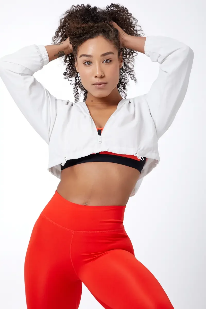 Model portfolio development session, commercial athletic look, orange sports bra with black underband, high-waist orange leggings, white cropped zip jacket open, three-quarter, both hands in hair, direct gaze, white background