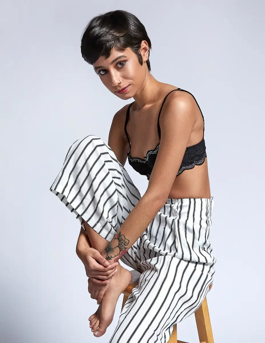 Eloissa, three quarter gaze, one leg tucked, white and black striped wide-leg trousers and tan suede cropped jacket, black lace bralette, barefoot, studio stool, East Williamsburg studio test shoot, makeup by Schanica Pickens, photographed by Scott P