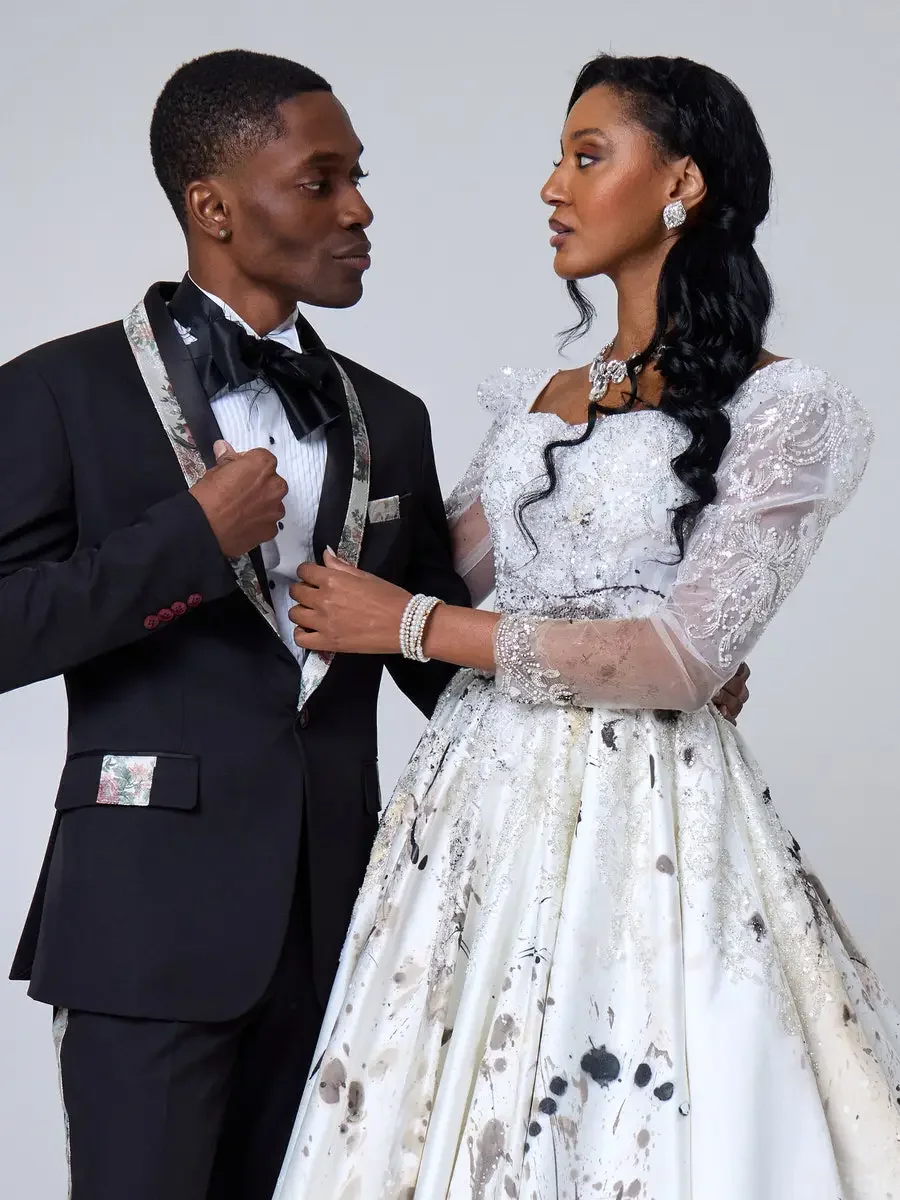 JUS10H 2025 New York Bridal Fashion Week, black tuxedo with floral-print shawl lapels, black satin bow tie, and matching floral pocket trim alongside white beaded paint-splatter ball gown with sweetheart neckline, sheer embroidered puff sleeves, and 