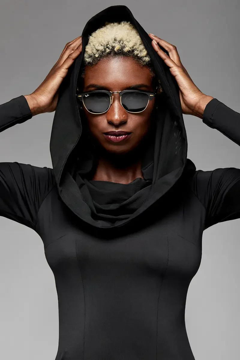Minika Ko lookbook, detail, black long-sleeve mini dress with oversized cowl collar, close crop showing hood framing face, dark sunglasses, smooth synthetic fabric, photographed by Scott Parker Photo