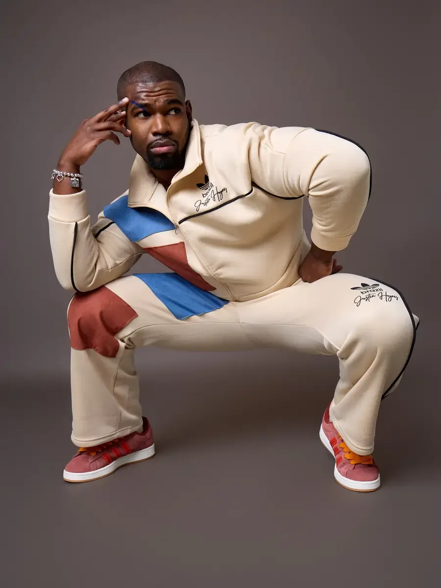 JUS10H x Adidas Brasil campaign, model in deep squat with hand raised to temple, cream tracksuit with diagonal blue and terracotta suede panels and black piping, Adidas Campus 00s in pink suede with red stripes and orange laces, warm brown background