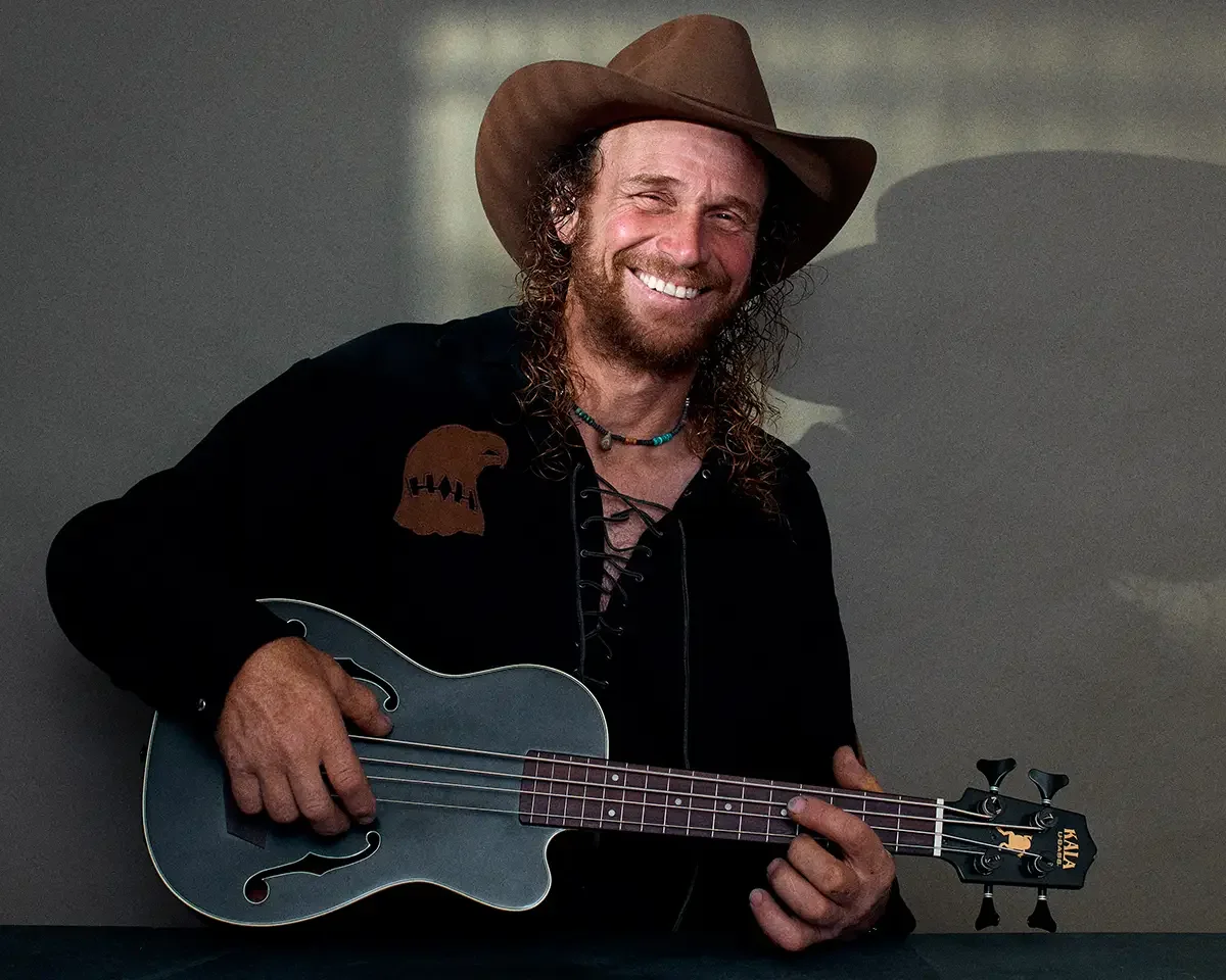 arborist, tree service, dan, playing bass uke, after work chill, branding portrait on muted army greenbackground window light, man wearing black shirt with lace up front and a brown cowboy hat, curly hair, smiling, scott parker photo