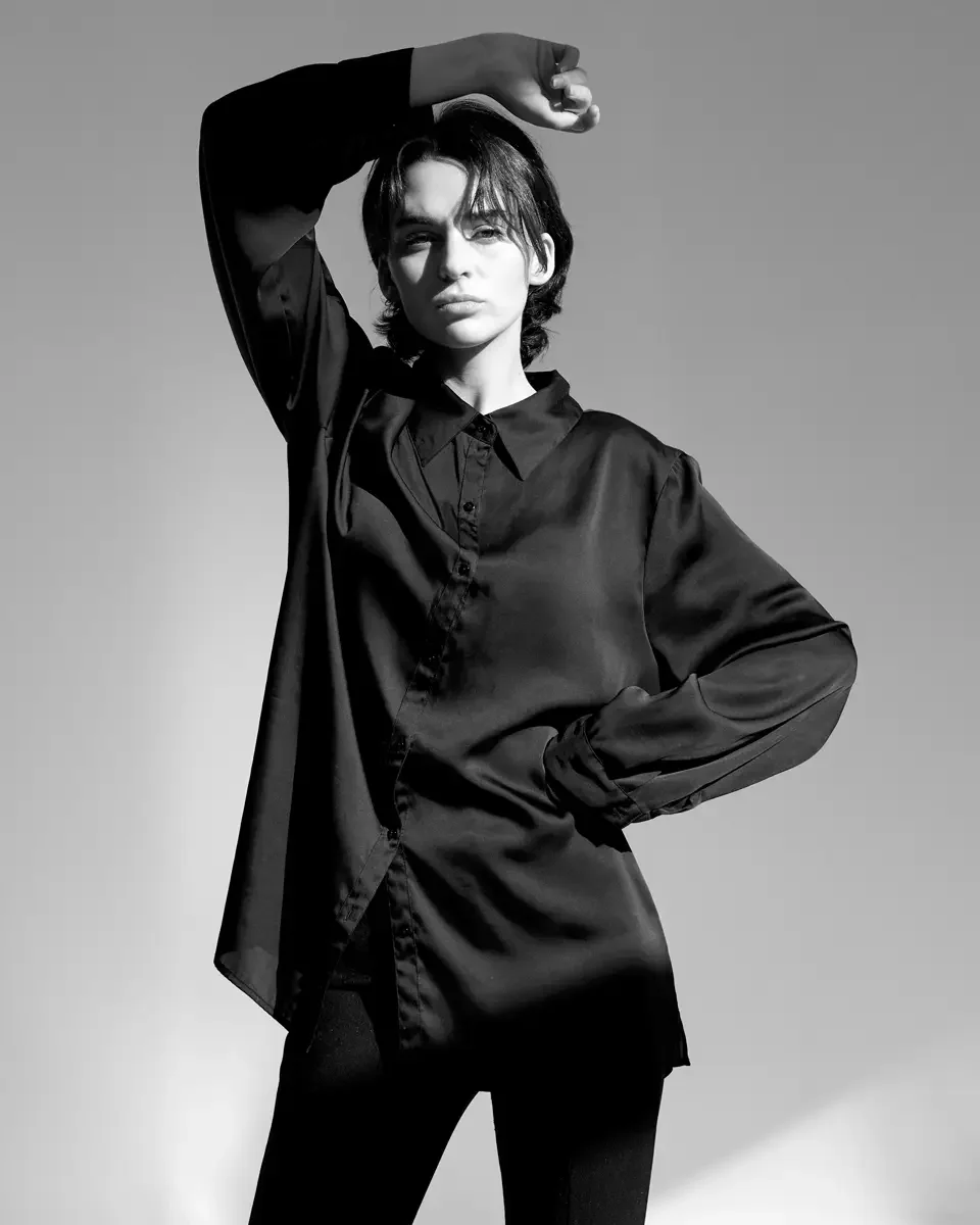 Tess, half-length, arm raised overhead with hand folded at wrist, opposite hand on hip, oversized black satin longline button-front shirt, black trousers, black and white, New York studio test shoot, makeup by Schanica, photographed by Scott Parker P
