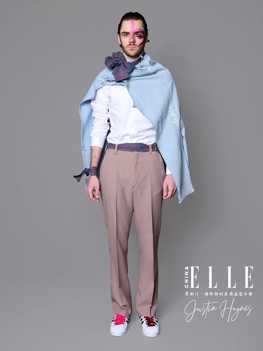 A male model with half pink face paint, wearing a white shirt, plaid pants, a denim jacket draped over shoulders, with a scarf and accessories, standing against a gray background, for ELLE China magazine, featuring Justin Haynes.