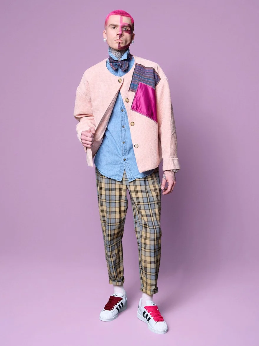 Person with pink hair and facial makeup, wearing a pink and multicolored patchwork jacket over a blue shirt, plaid pants, and white sneakers with red and black stripes, standing against a light purple background.