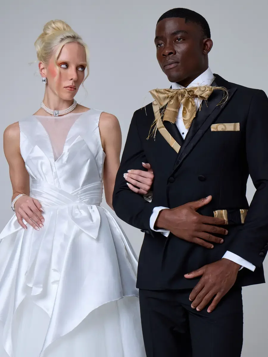 JUS10H 2025 New York Bridal Fashion Week, white sculptural ball gown with illusion V-neckline, oversized bow waist, and asymmetric peplum alongside black double-breasted tuxedo with gold jacquard bow tie with feather detail and matching pocket square