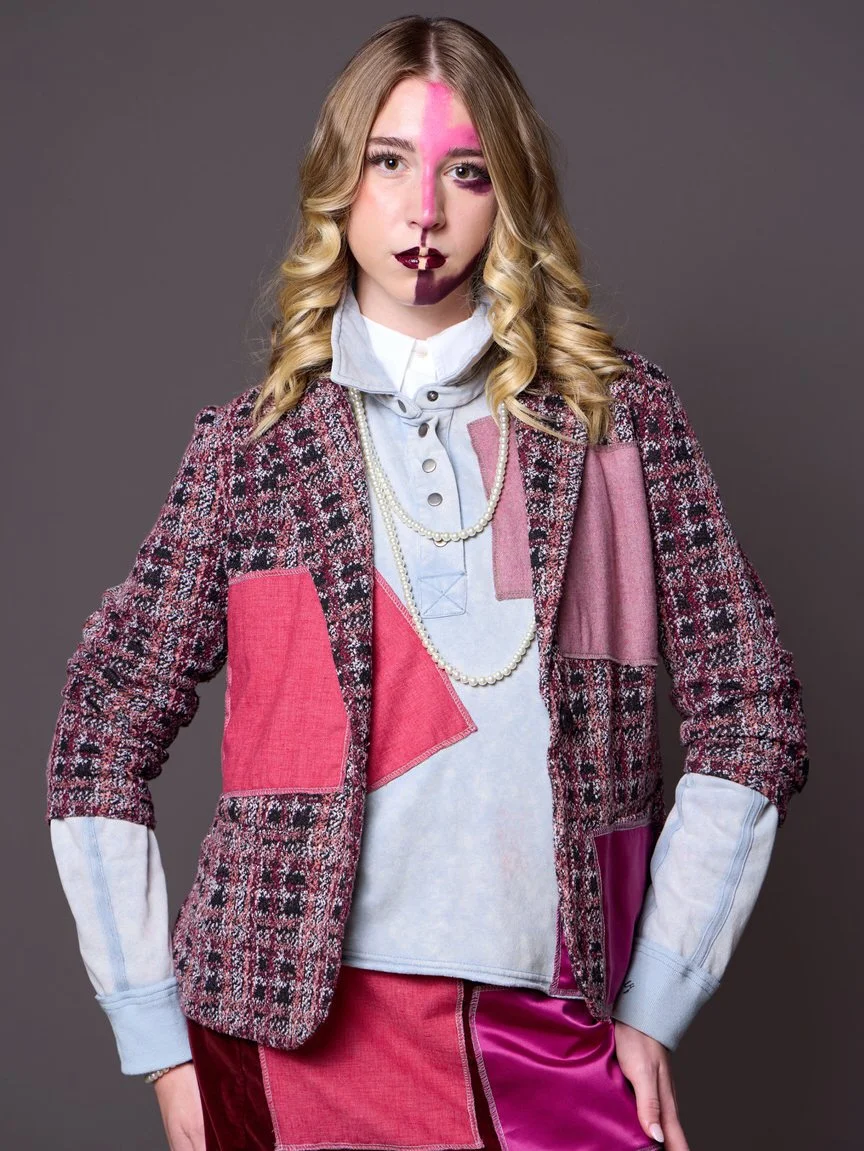 A woman with blonde curly hair and painted face with pink and dark purple designs, wearing a colorful patchwork jacket, a white collared shirt, layered pearl necklaces, and a pink and purple skirt, posing against a gray background.