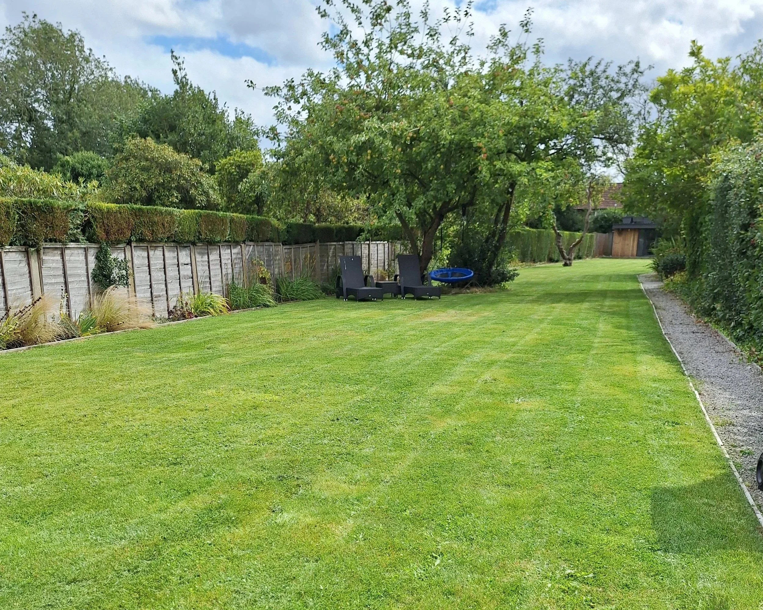 A well-maintained backyard with a large green lawn, surrounded by trees, shrubs, and a wooden fence.