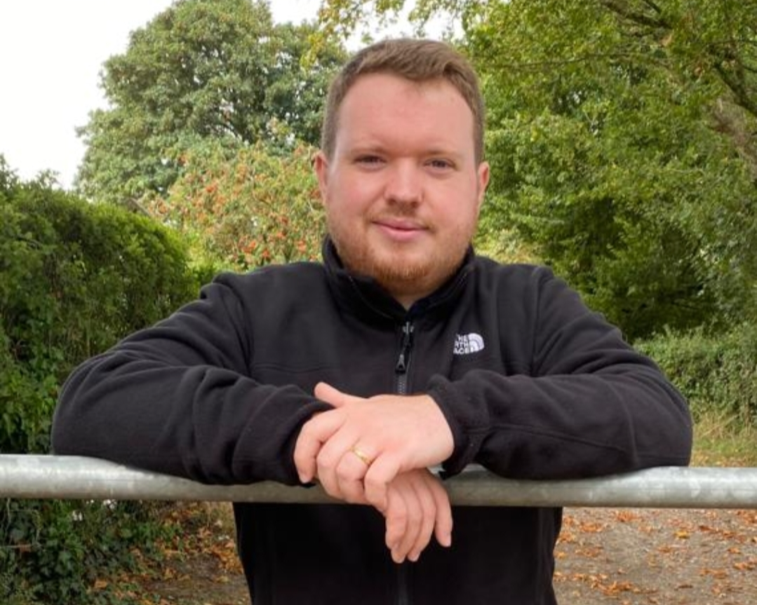 A man with short brown hair and a beard outdoors, leaning on a metal railing, wearing a black North Face jacket, with green trees and bushes in the background.