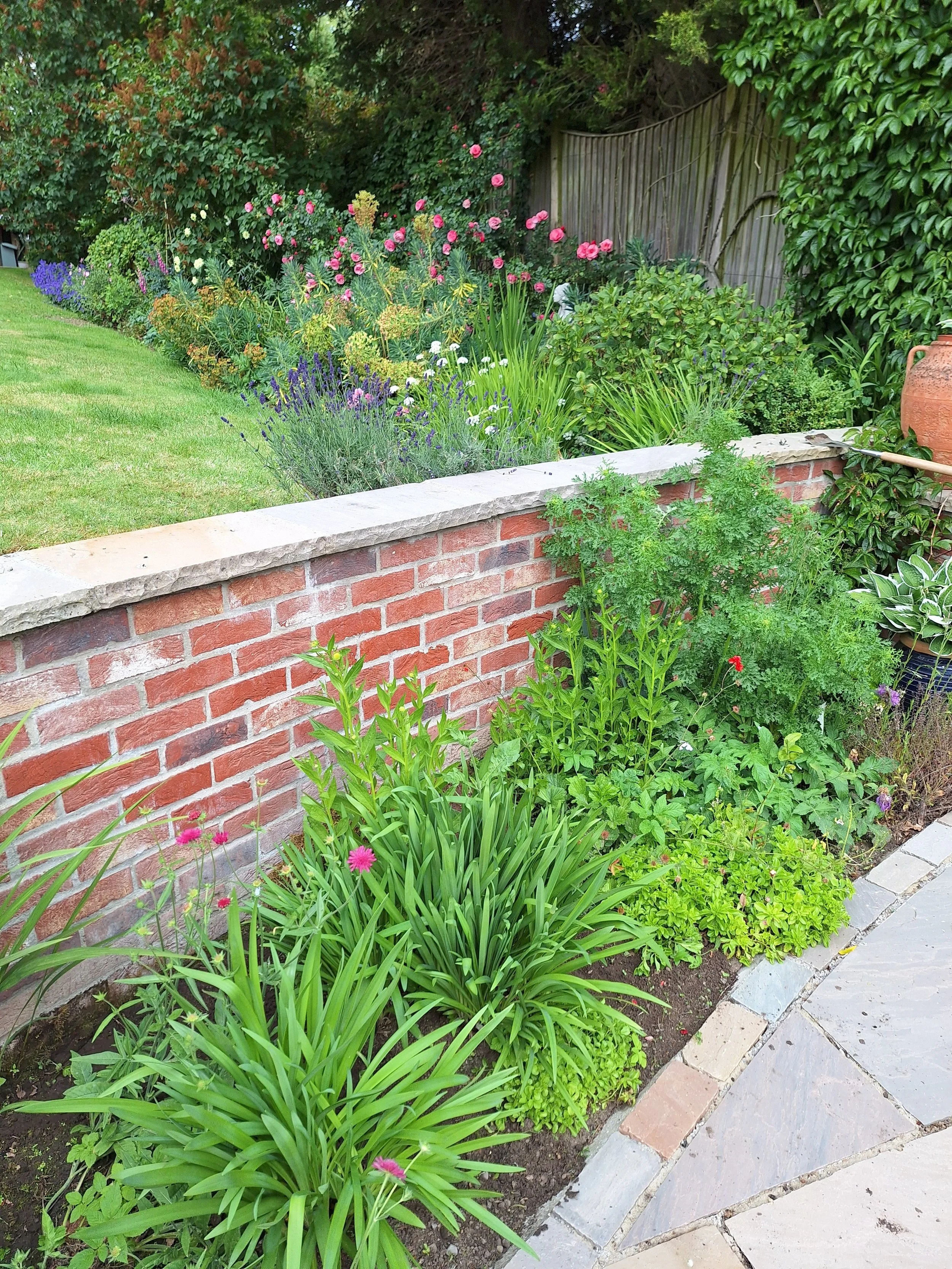 A garden with a brick wall and a variety of colorful flowers and green plants, next to a stone pathway.