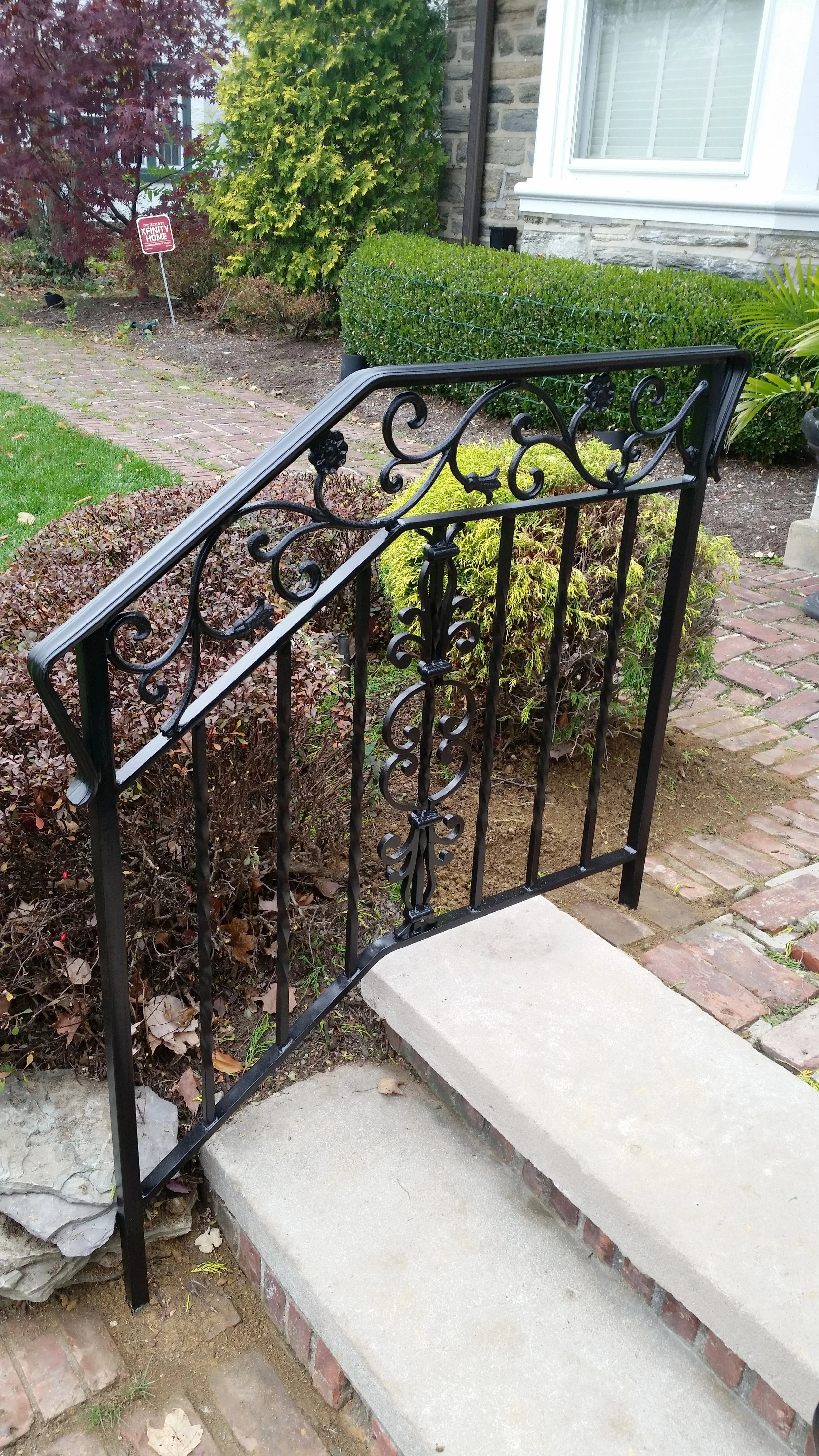 Black wrought iron railing on a small concrete step leading to a garden pathway in front of a house. The garden has shrubs, a small tree, and a lawn with a sign in the background.