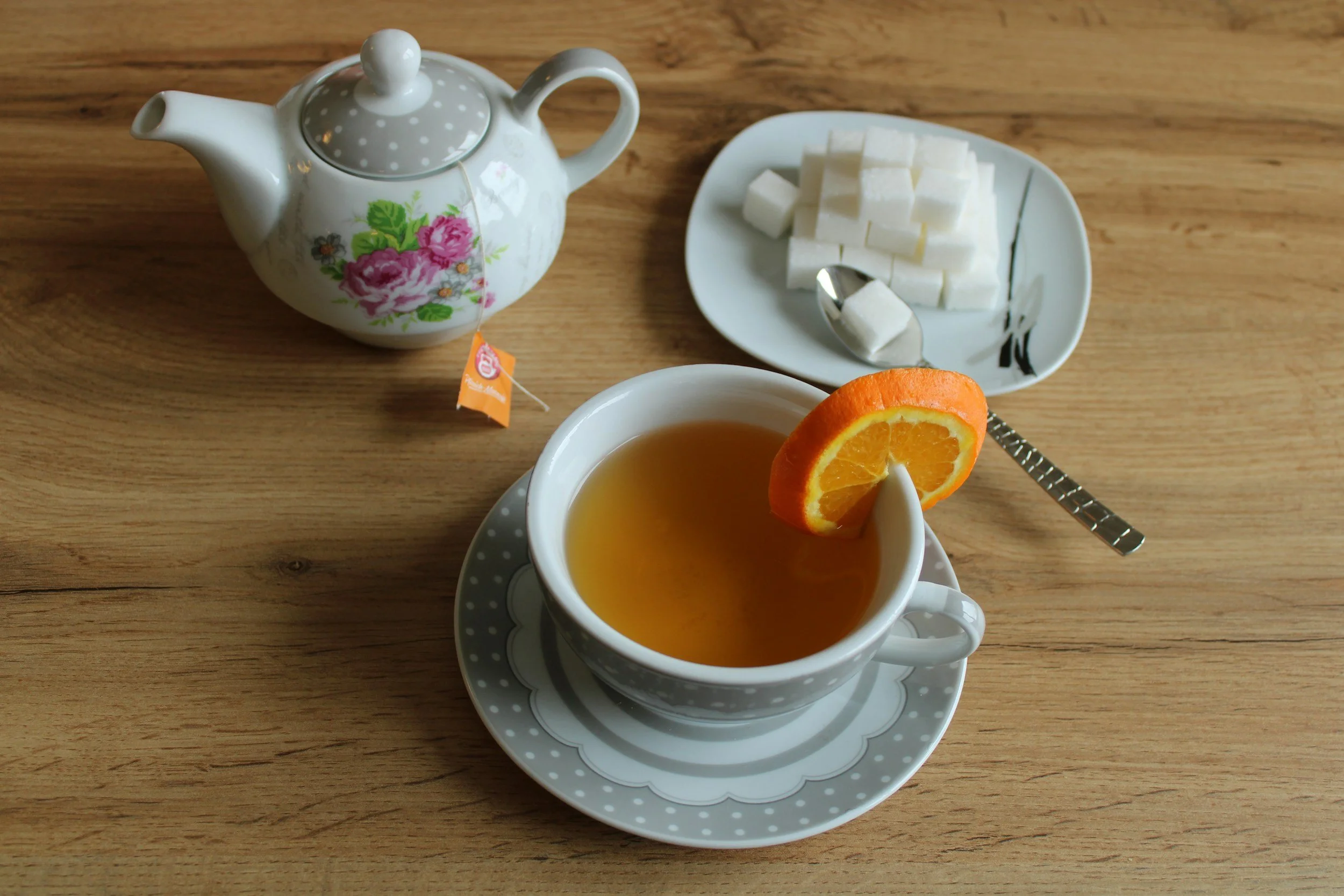 A cup of tea with an orange slice, a teapot with floral design, a small plate of sugar cubes, and a teaspoon on a wooden table.