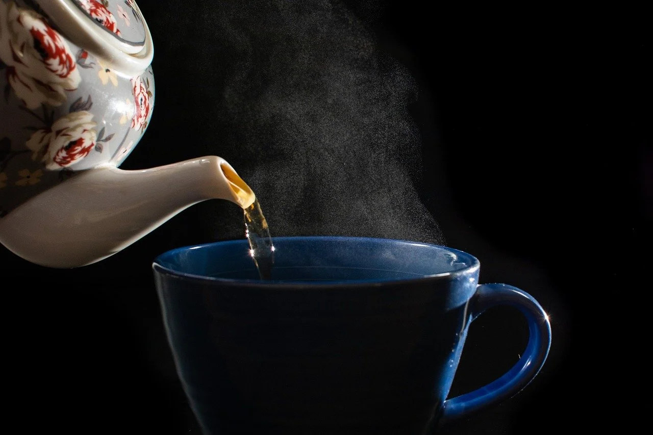 A porcelain teapot pouring tea into a blue cup with steam rising against a black background.