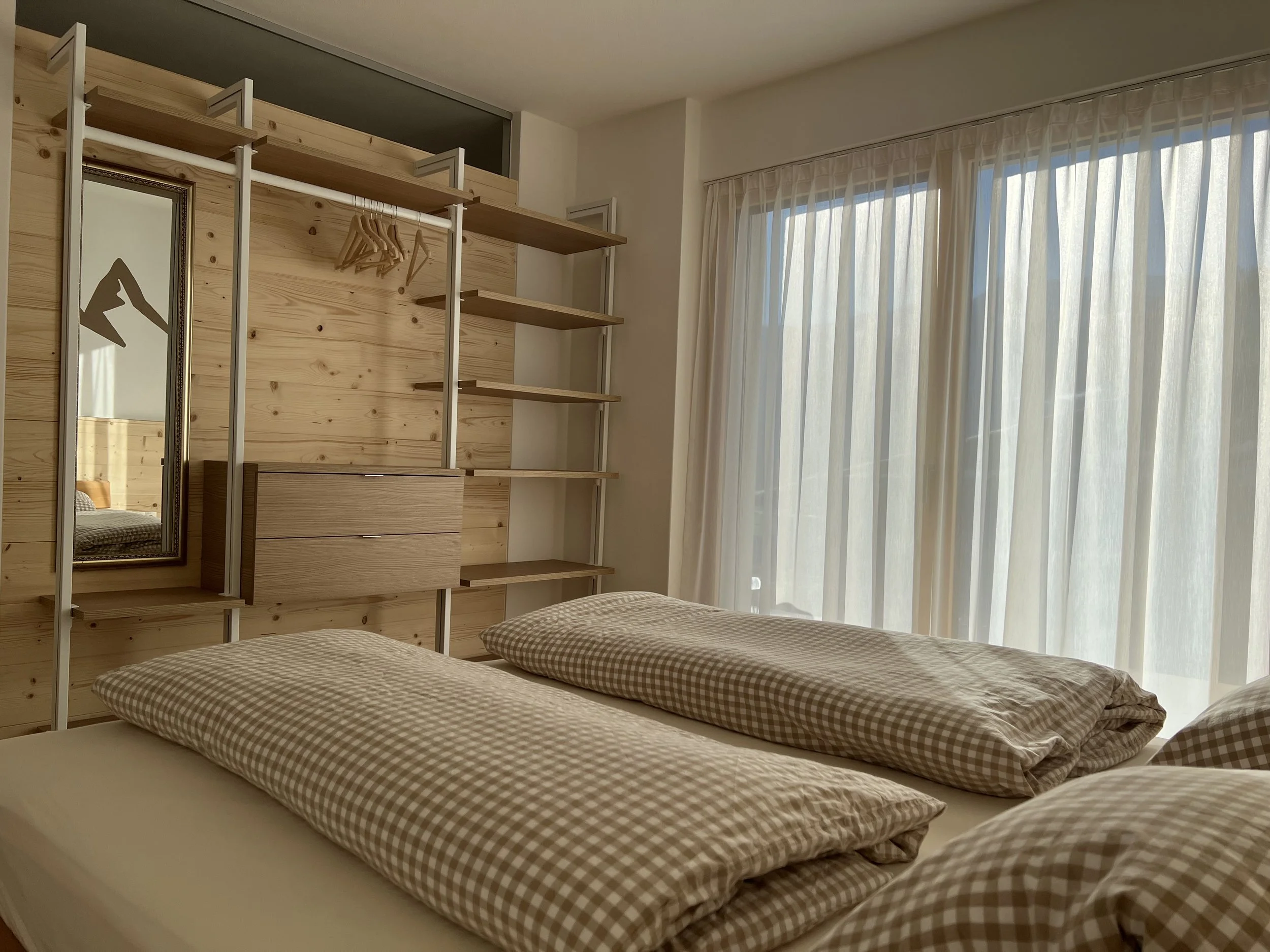 Schlafzimmer mit zwei Betten, natürlichen Holzmöbeln, eine große Fenster mit weißen Vorhängen, Spiegel, offene Regale und eine kleiderstange mit Kleiderbügel.