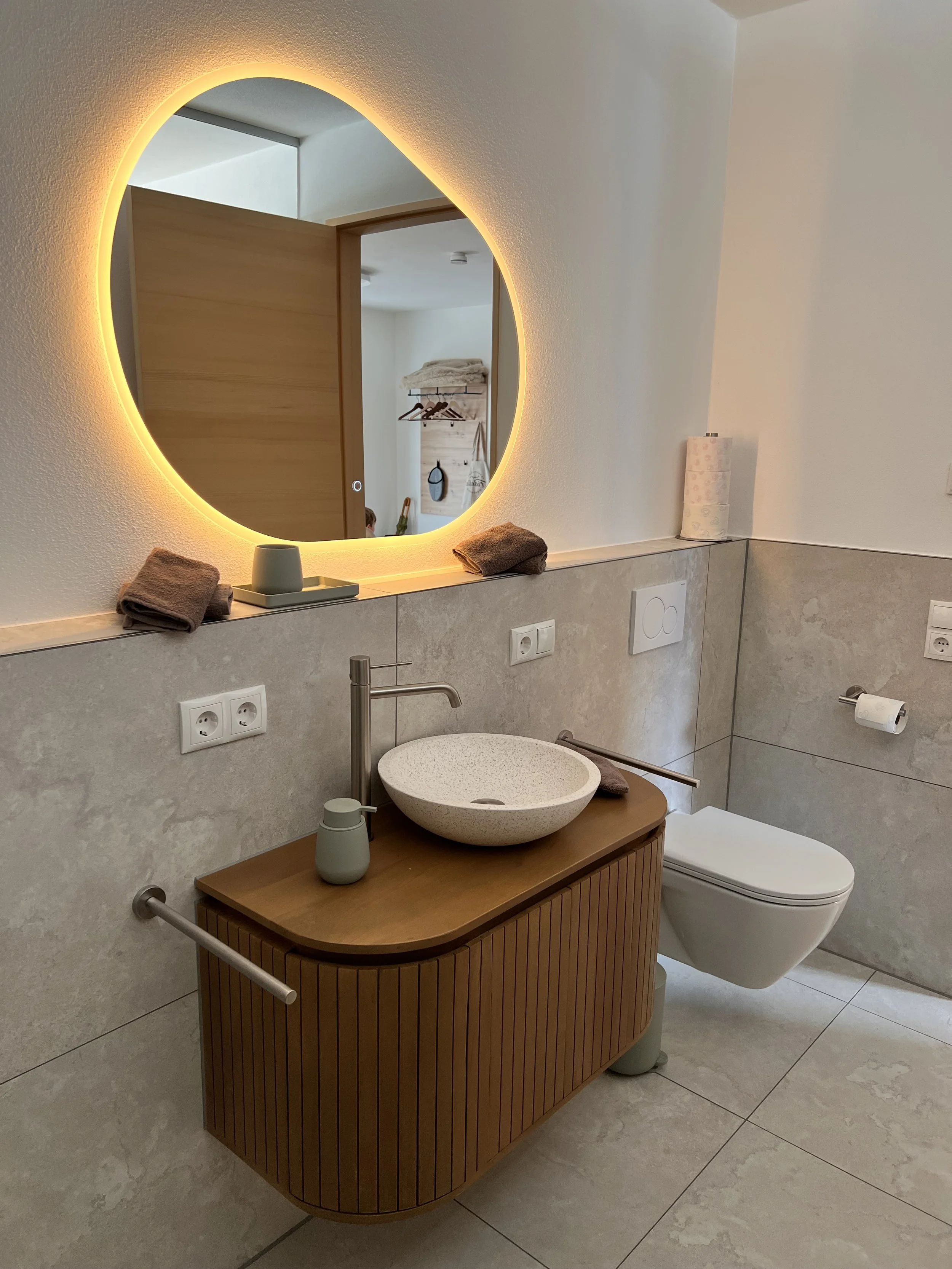 Moderner Badezimmerbereich mit einem abgerundeten Waschtisch, einer runden Pflanzen- oder Handtuchschale auf Holzoberfläche, einem Wandspiegel mit LED-Beleuchtung, einer Toilette, Handtüchern und Papierrollen.