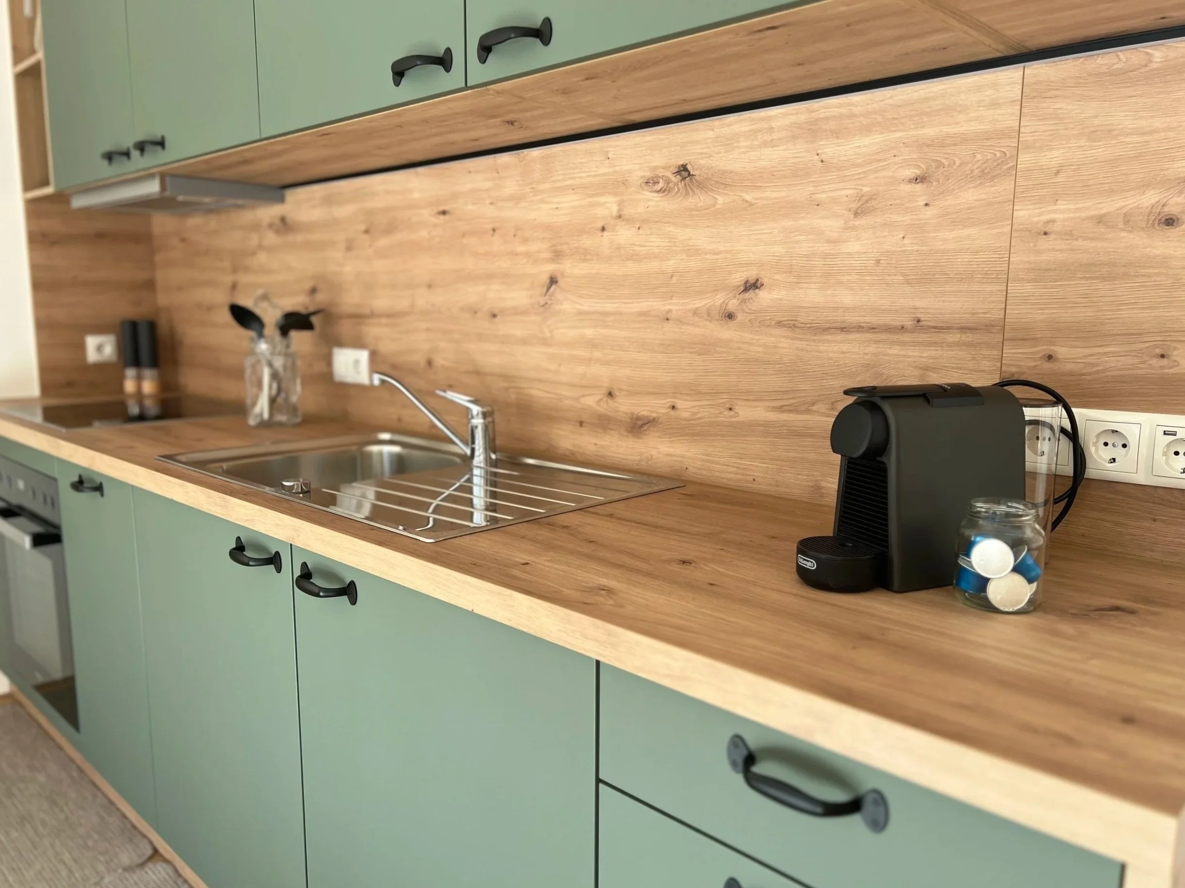 Küchenarbeitsplatte mit Wasserkocher, Kaffeekapseln, Spüle, Werkzeug und dekorative Flasche mit Zweigen, grüne Küchenschränke, Holzarbeitsfläche und Holzrückwand.