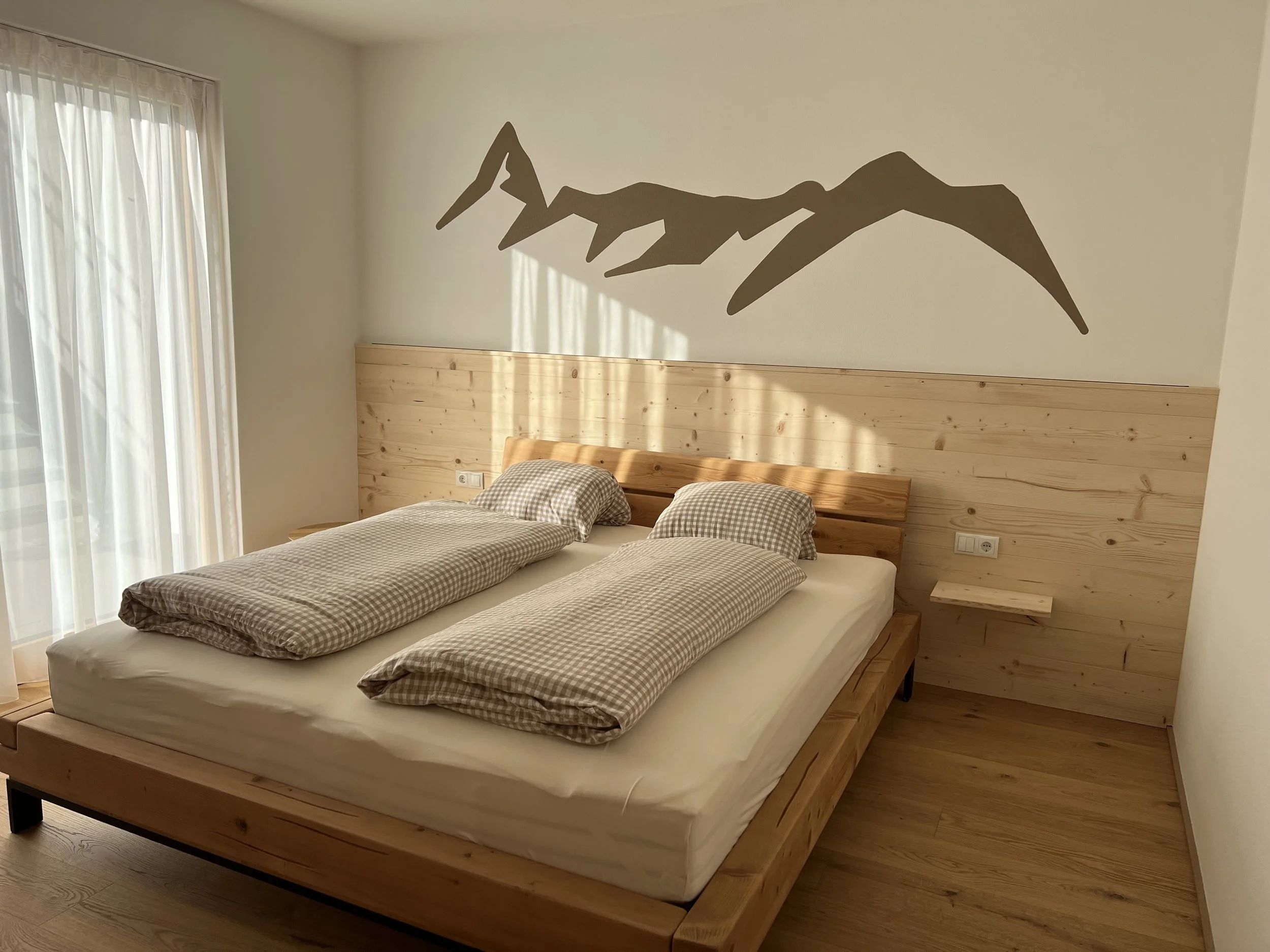 Ein Schlafzimmer mit einem Doppelbett, Holzpaneele an der Wand, zwei Kissen, Bettdecke im Karo-Muster, Vorhänge vor einem Fenster, eine Wanddekoration in Form einer Bergsilhouette.