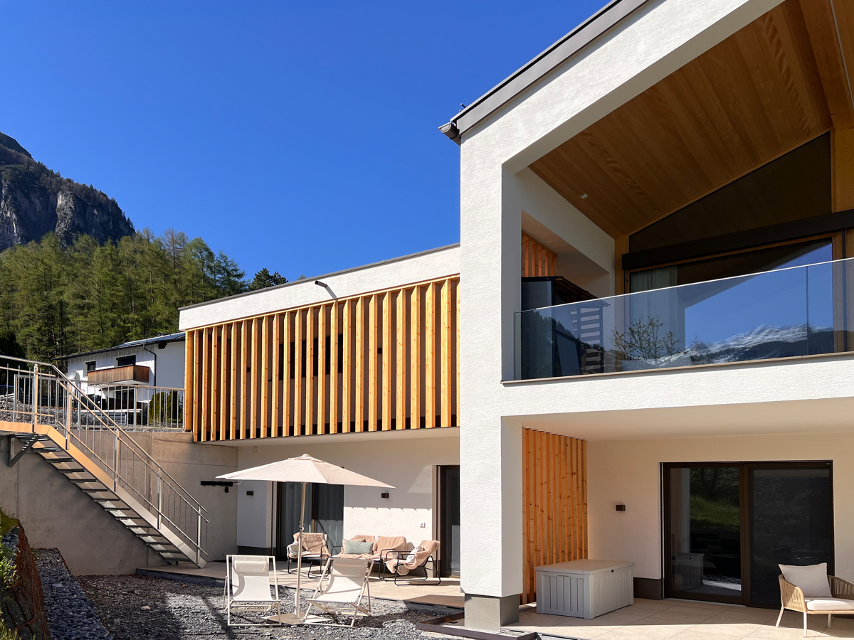 Modernes zweistöckiges Haus mit Balkon, Terrasse und Holzverkleidungen, umgeben von Bergen und Bäumen.