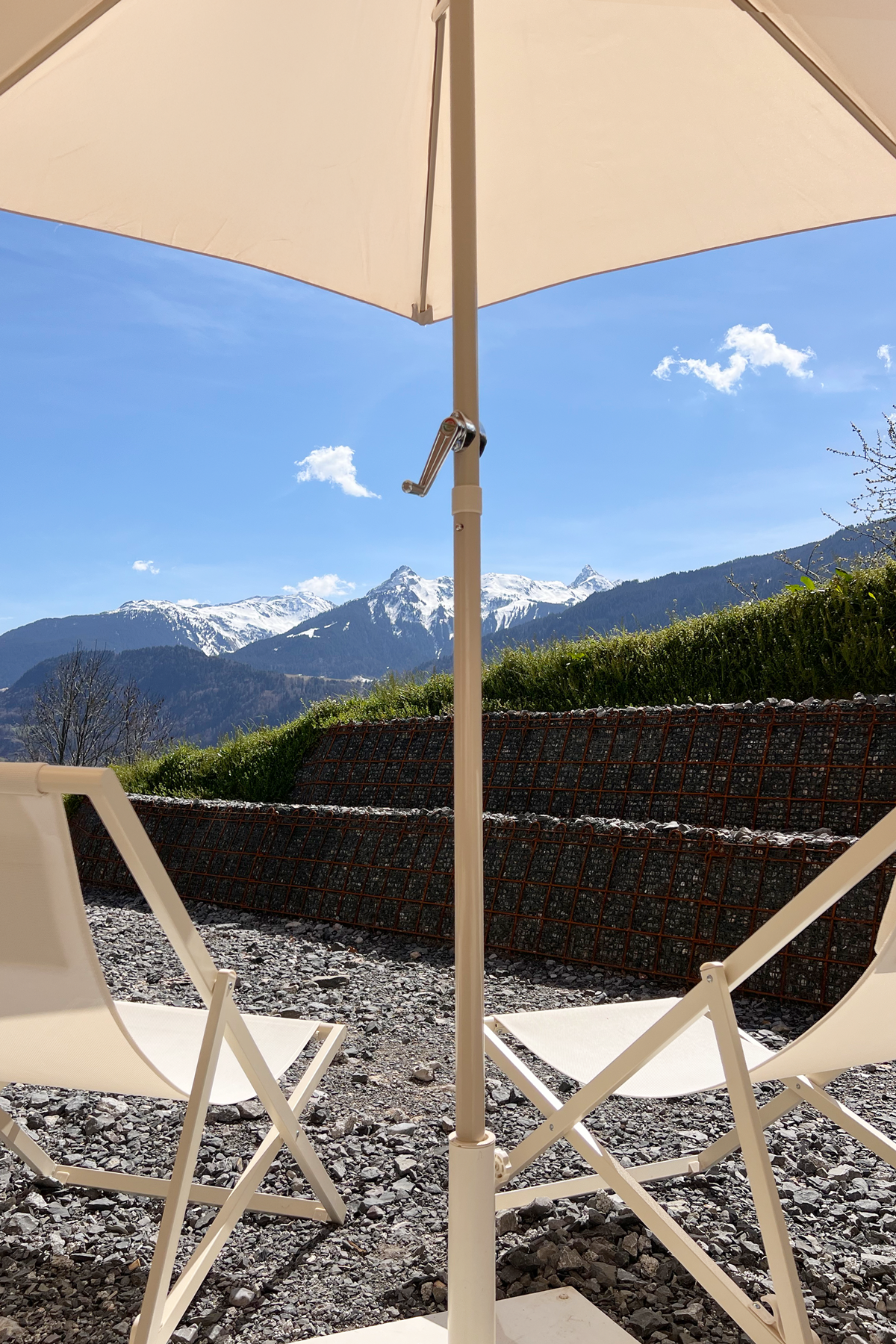 Blick auf Berge mit schneebedeckten Gipfeln, Sicht von einem Garten mit weißem Sonnenschirm und zwei weißen Liegestühlen auf kiesbedecktem Boden.