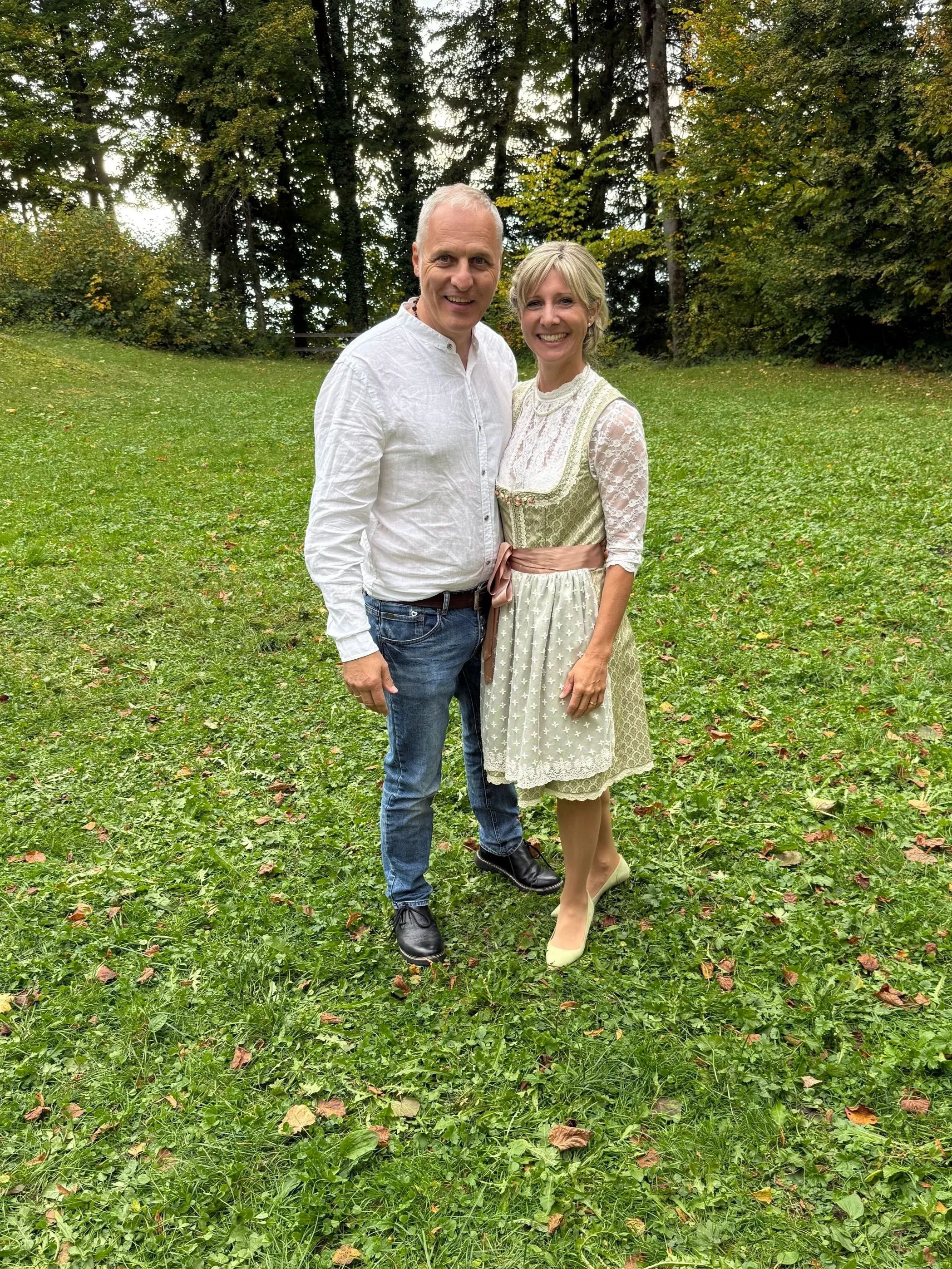 Ein Mann und eine Frau stehen in einem grünen Park, beide lächeln und betrachten die Kamera. Der Mann trägt ein weißes Hemd und Jeans, die Frau trägt ein traditionelles Dirndl. Im Hintergrund sind Bäume und eine bewaldete Umgebung sichtbar.