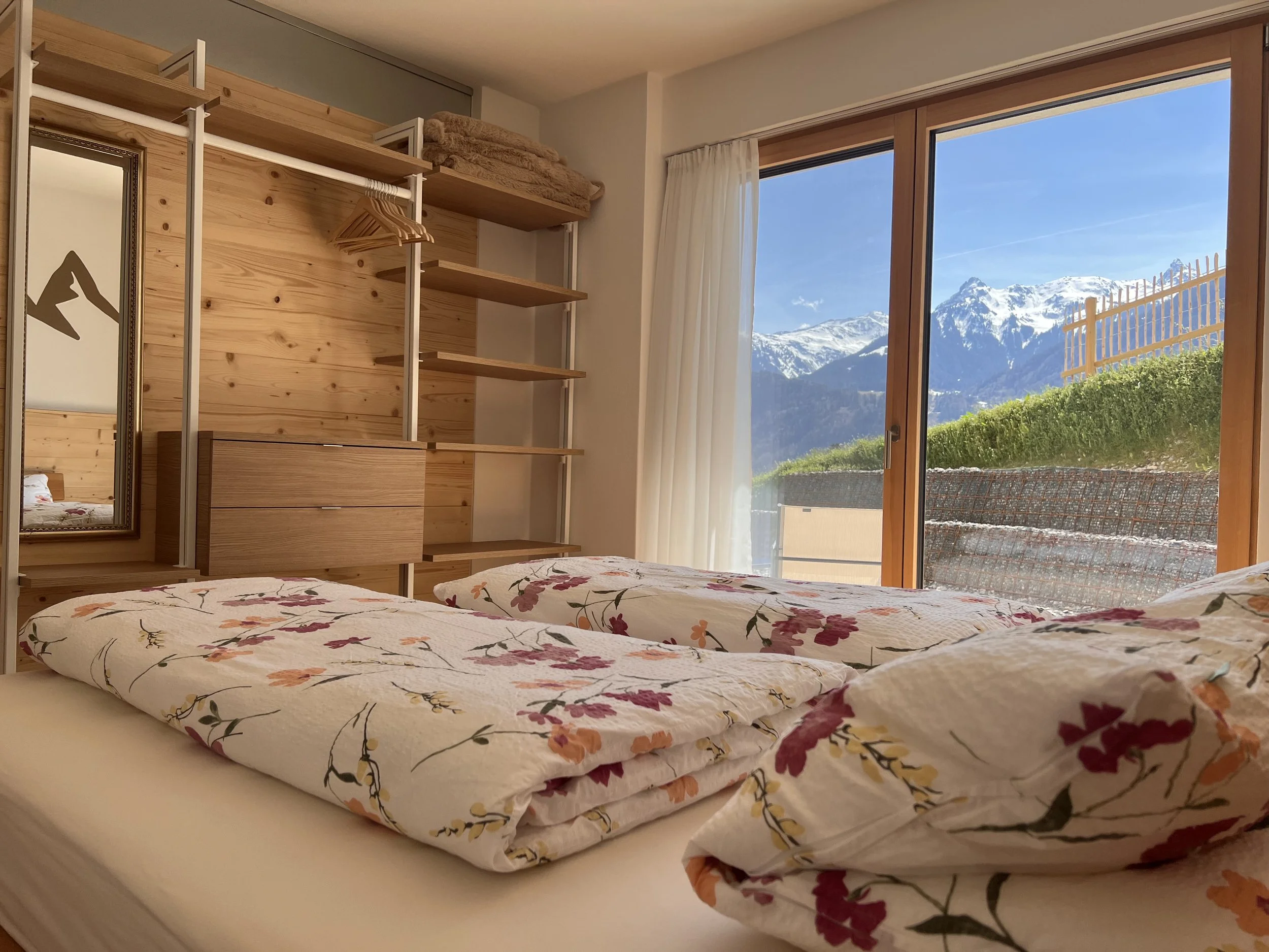 Schlafzimmer mit zwei Betten, Berge im Hintergrund, große Fenster, Gardinen, Holz-Dekoration.
