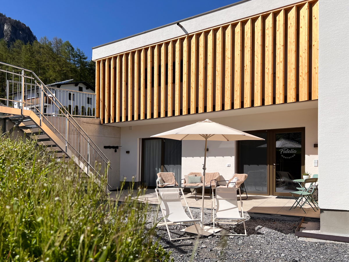 Moderner Privatwohnsitz mit Terrasse, Sonnenschirm, Gartenmöbeln, Holzverkleidung außen, umliegende Natur, blauer Himmel.