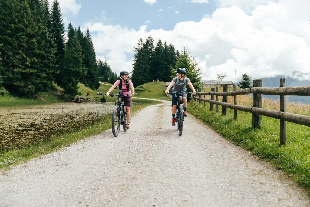 Zwei Mädchen mit Helmen, die auf einem Fahrradweg in der Natur Fahrrad fahren, umgeben von Bäumen und Bergen.