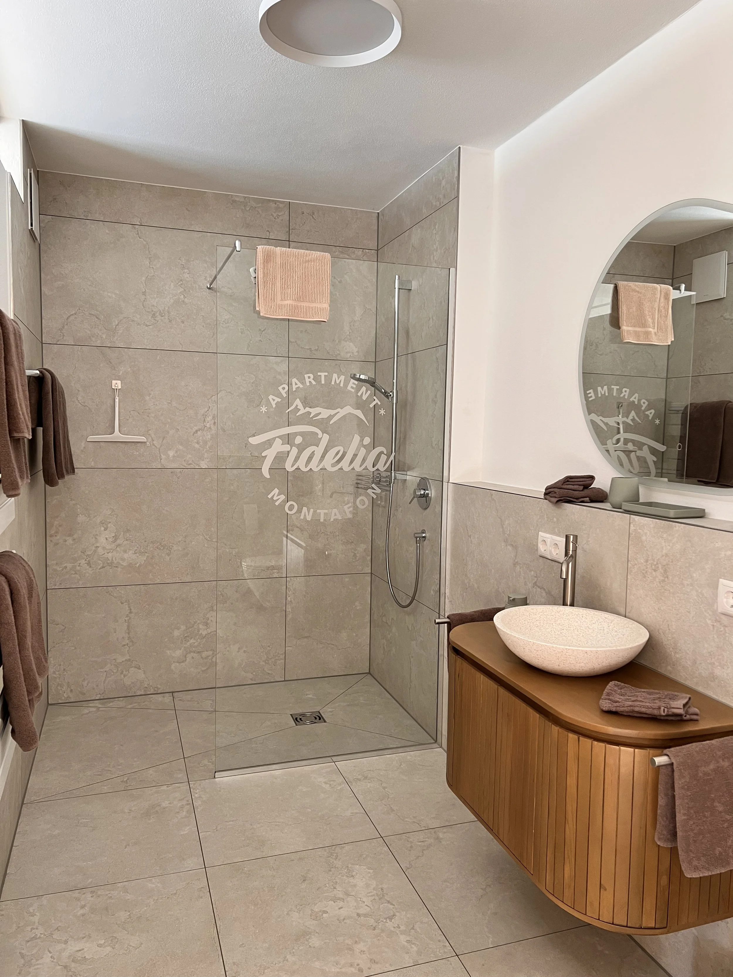 Modernes Badezimmer mit beigem Fliesen, offener Dusche, runder Waschschale auf einem Holzunterschrank, Spiegel, Handtüchern in Braun und Beige.
