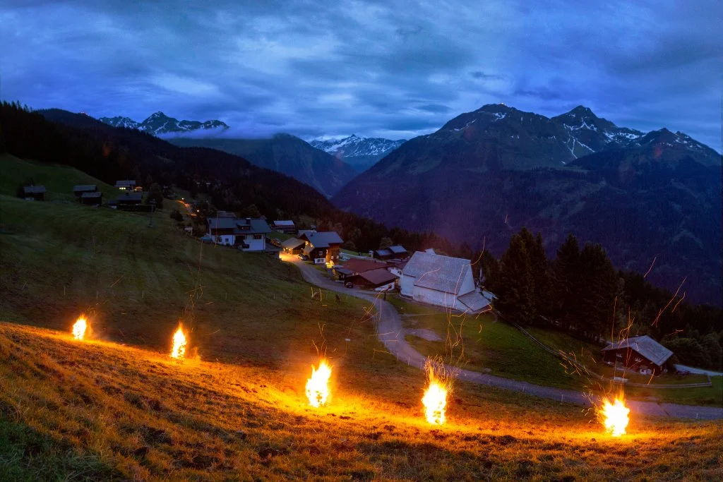 Abendliche Berglandschaft mit kleinen Häusern, leuchtenden Feuerstellen am Hang und Bergen im Hintergrund.