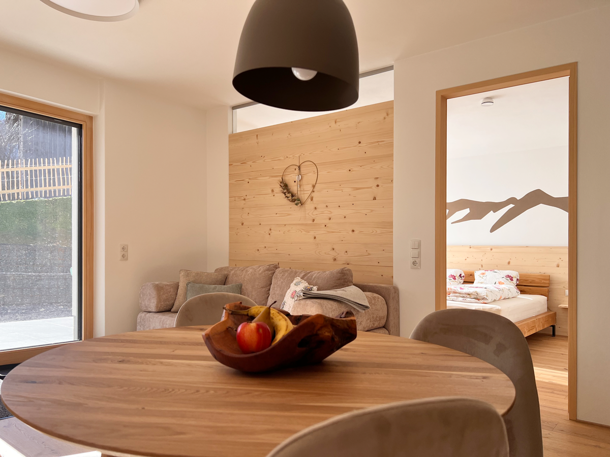 Innenansicht eines modernen, minimalistischen Wohnzimmers mit runden Holztisch, Obstschale und Blick in Schlafzimmer mit Wandkunst.