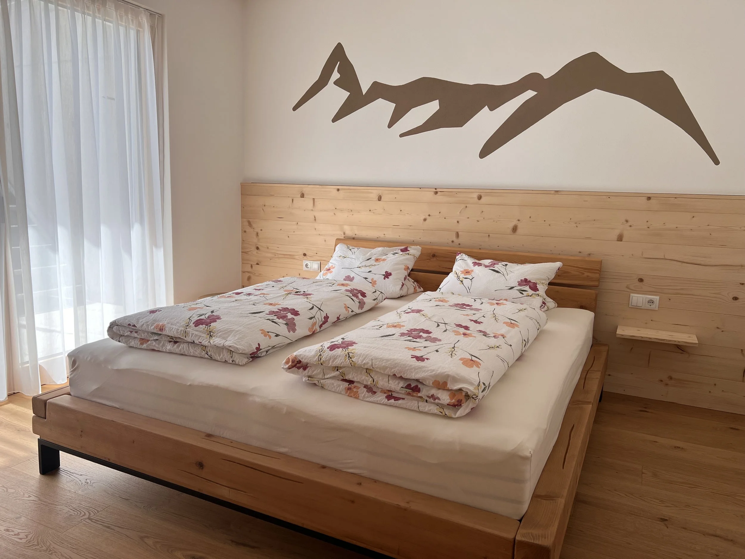 Ein Bett mit zwei Schlafsäcken, ein Holzkopfteil, Wanddeko in Form einer Berglandschaft, helle Vorhänge im natürlichen Licht.