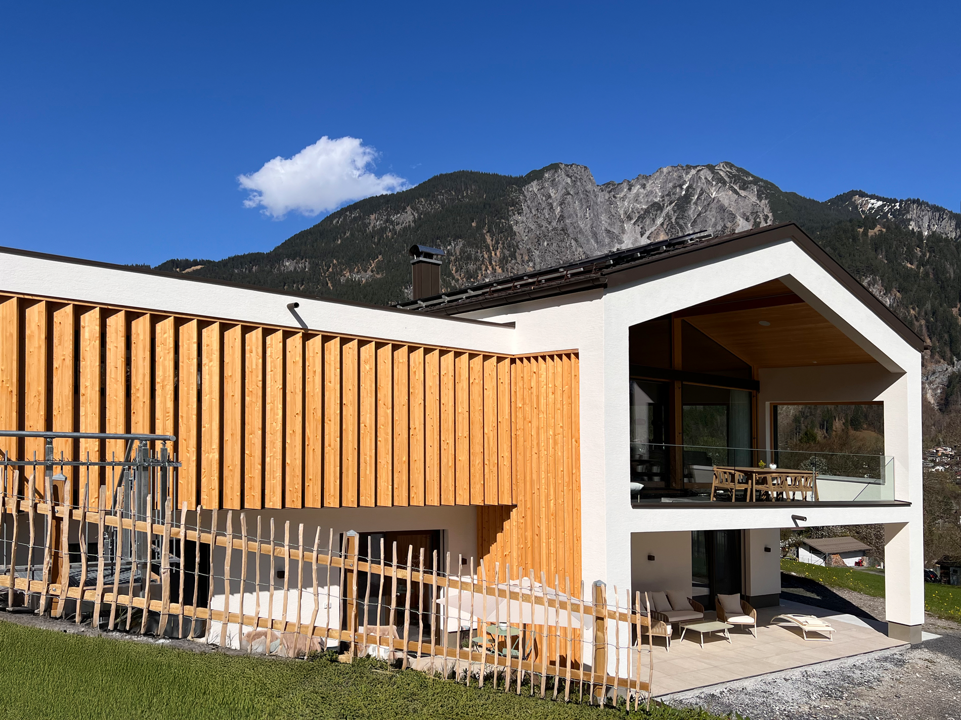 Modernes Haus mit Holzverkleidung, Balkon mit Tisch und Stühlen, davor Garten mit Liegen, Berge im Hintergrund, blauer Himmel