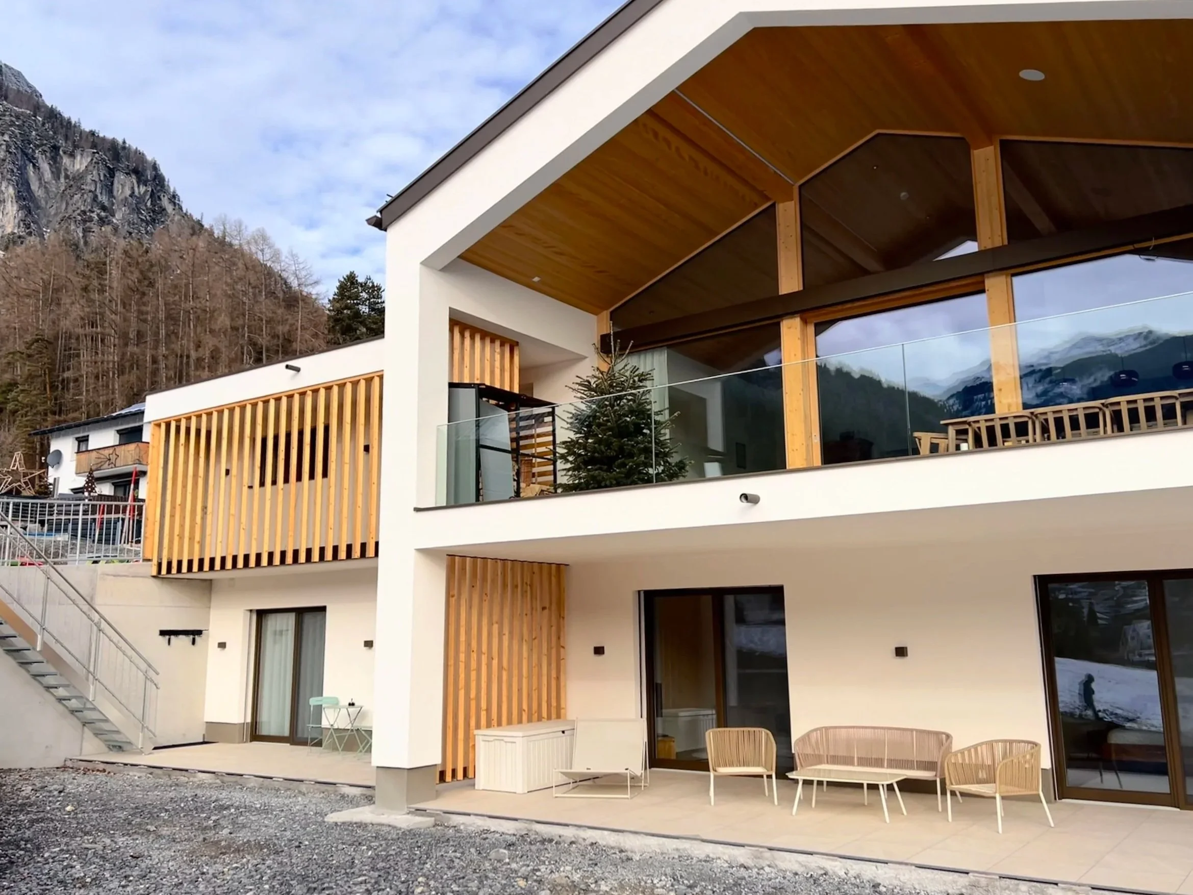 Modernes mehrstöckiges Haus mit weißen Wänden, viel Holzverkleidung und großen Fenstern, umgeben von Bergen und Bäumen, auf der Terrasse sind Gartenmöbel und eine Weihnachtsbaum