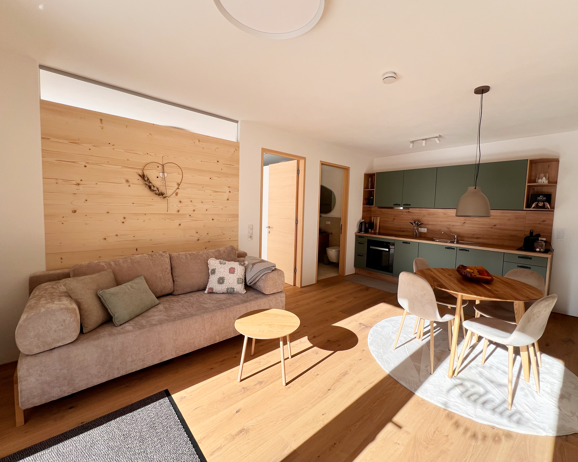 Offene Wohnküche mit Esstisch, Wohnzimmer mit Couch, Holzdeko und einem Herz an der Wand, Holzboden, durch Sonnenschein beleuchtete Raum.