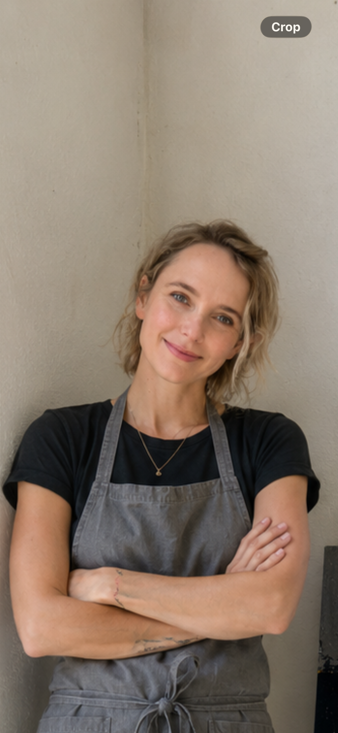 A woman with wavy blonde hair, wearing a black t-shirt and gray apron, smiling with arms crossed, standing against a plain wall.