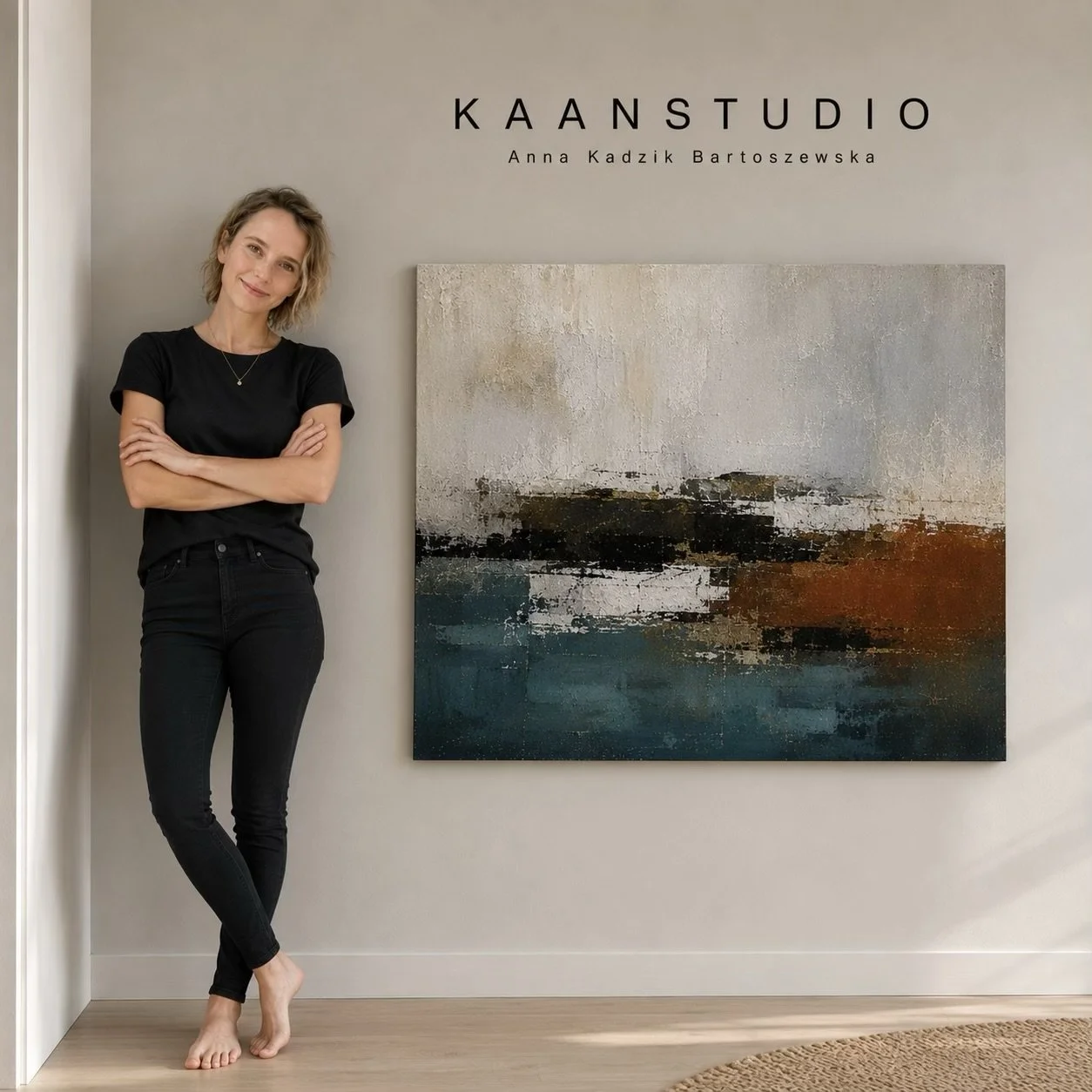 A woman with short curly blonde hair standing next to an abstract painting on a gallery wall, smiling with arms crossed, dressed in a black t-shirt and black jeans, barefoot.