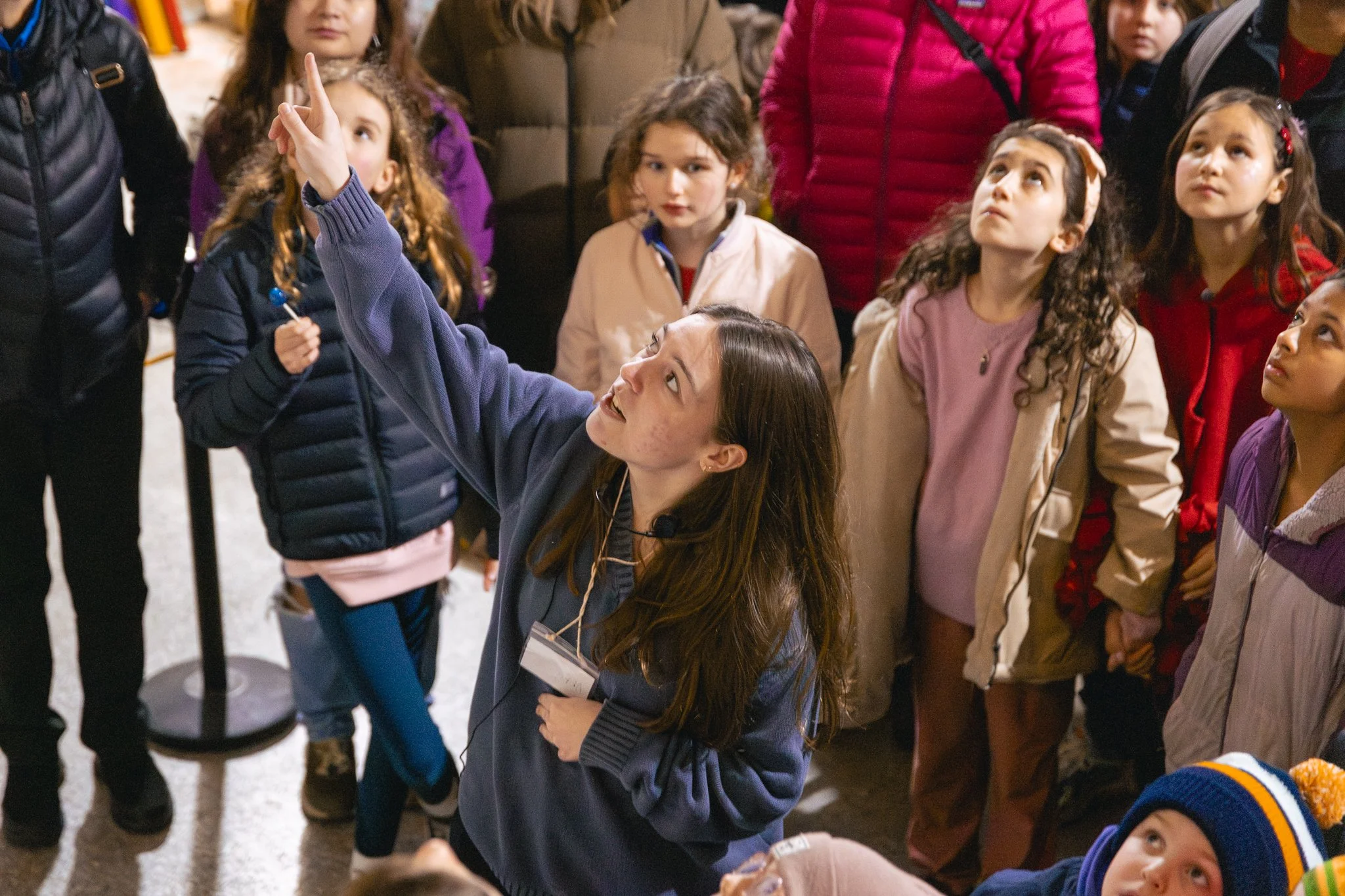 Children and adults gathered indoors, with one girl pointing upwards.
