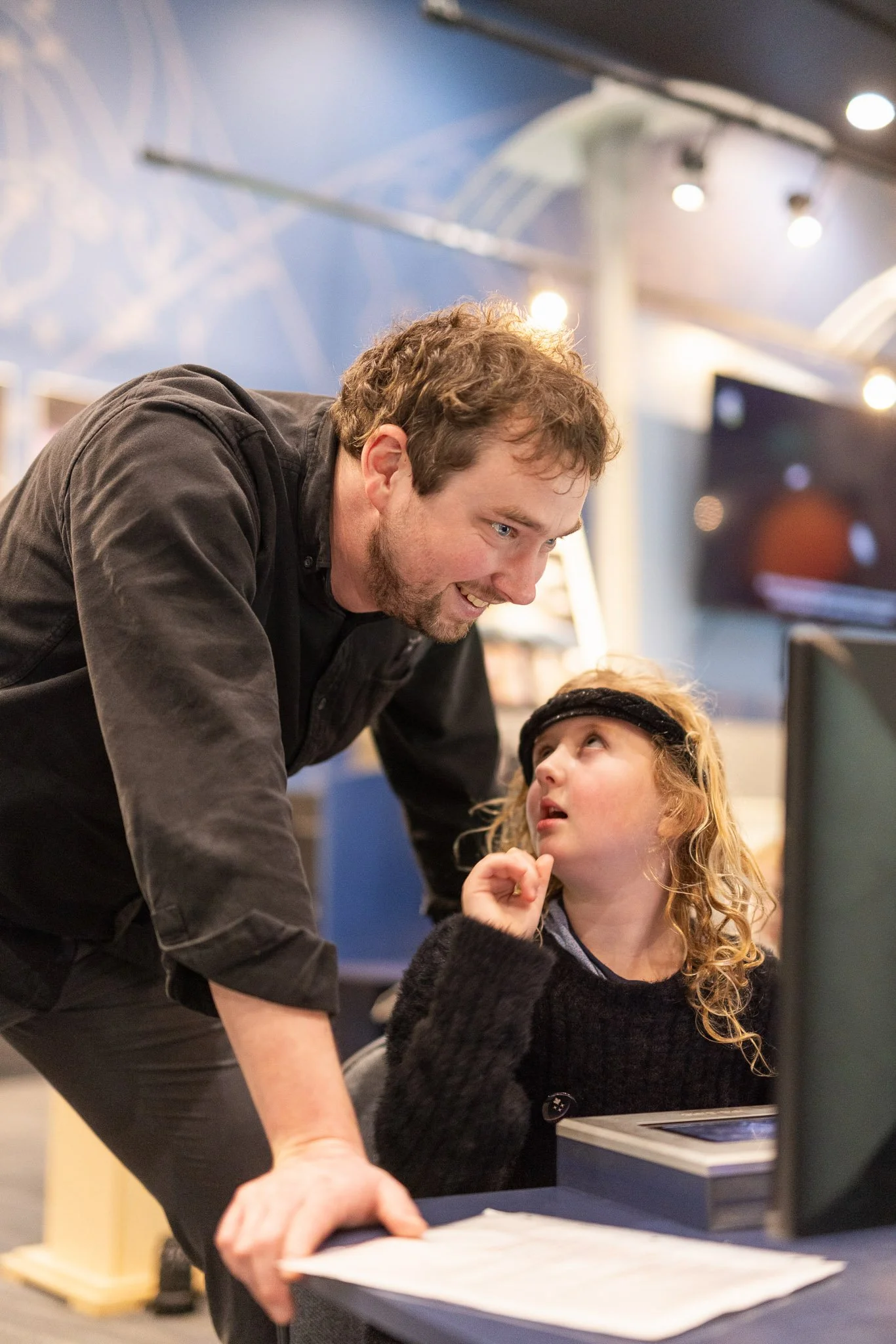 A man and a young girl looking at a computer screen in a store or technology setting, with warm indoor lighting and a TV in the background.