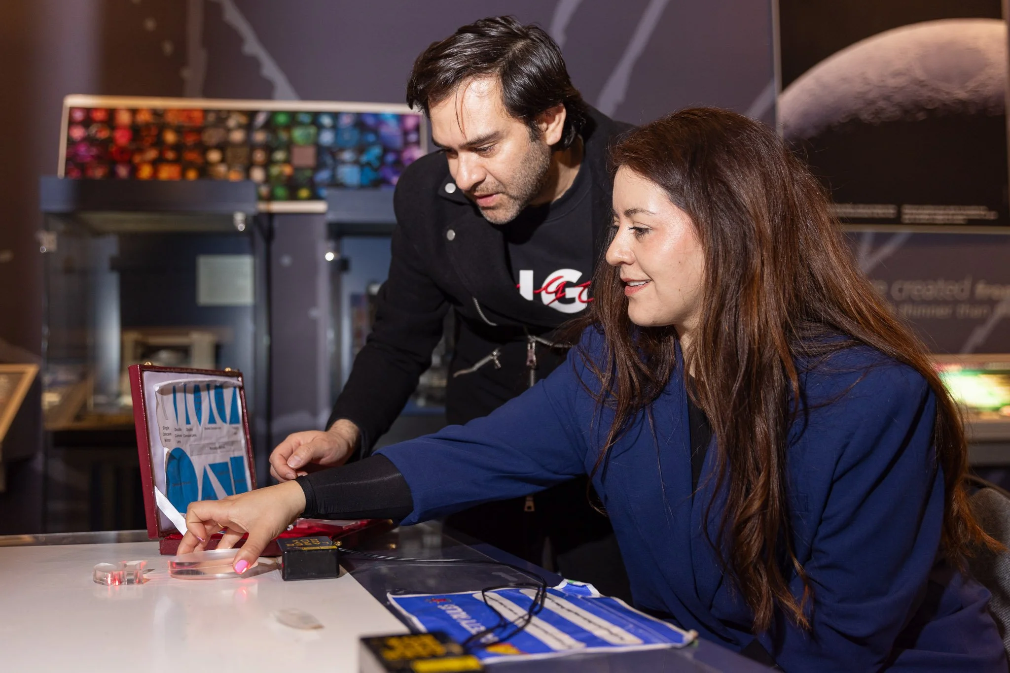 A woman in a blue jacket and a man with dark hair and beard are sitting at a table, conducting a scientific experiment with small transparent containers and a laser device. They are focused and engaged, with various scientific equipment and colorful 