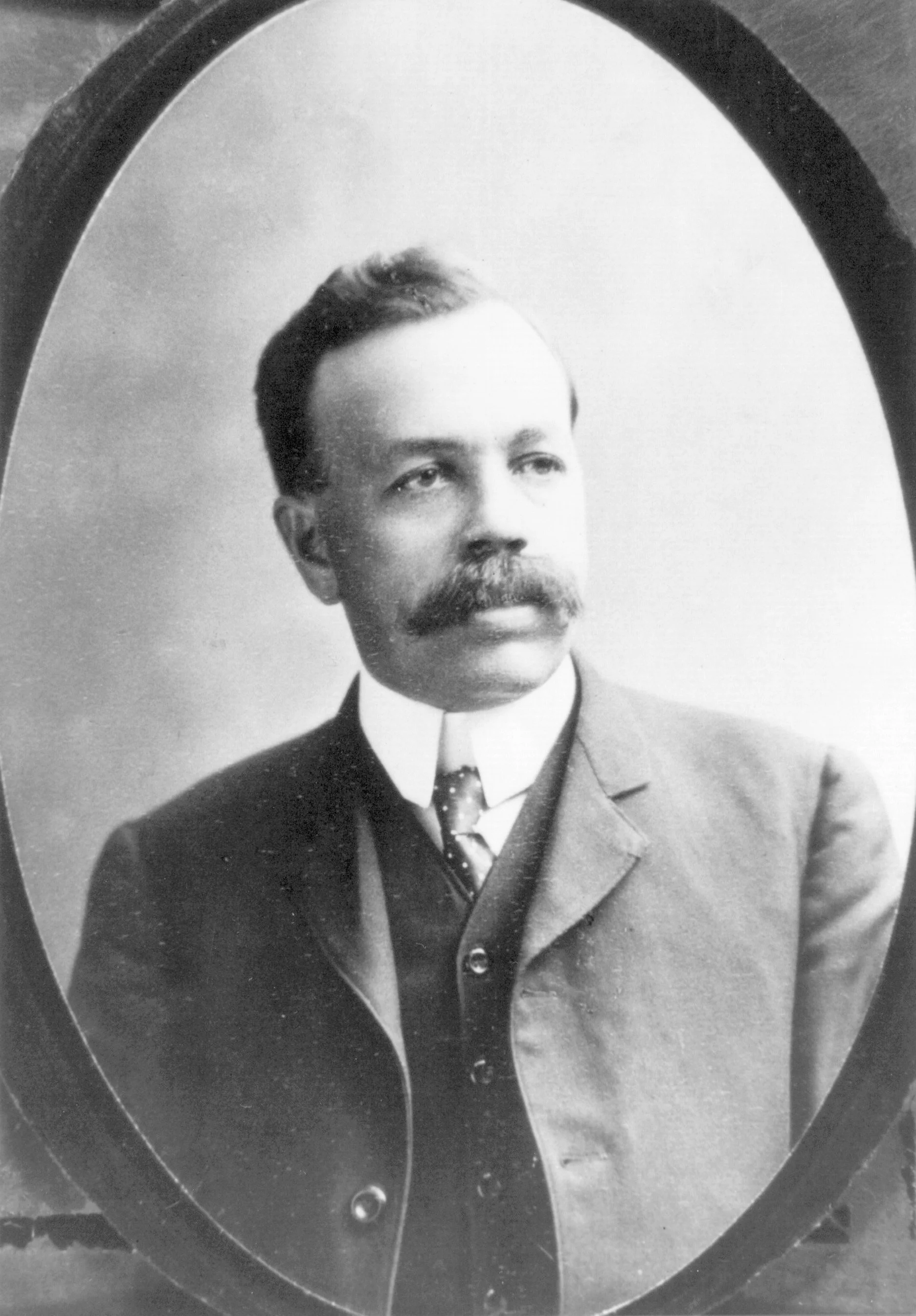 Black and white portrait of a man with a mustache, wearing a suit and tie, inside an oval frame.
