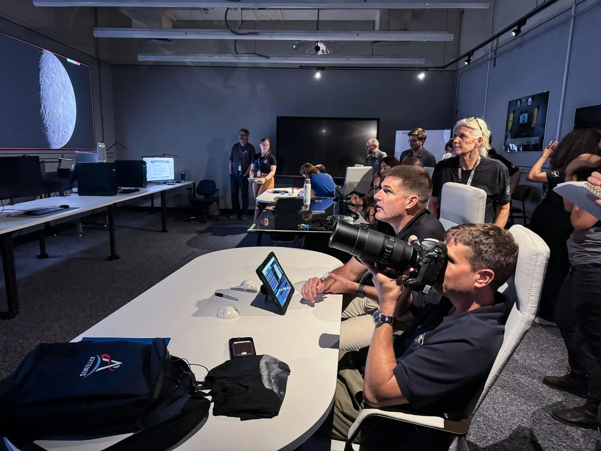 NASA scientists and technicians in a conference room with computers and monitors, preparing for a lunar mission, with a large lunar surface image displayed on a screen.