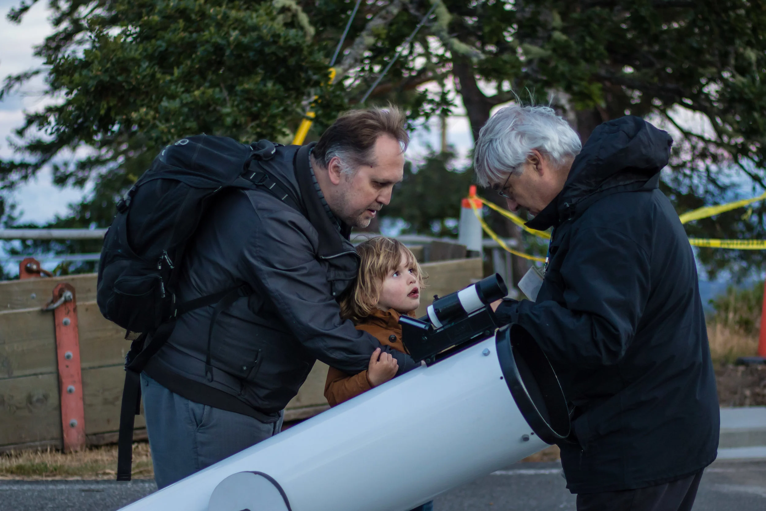 Two adult men and a young girl examining a telescope outdoors near trees and a wooden fence.