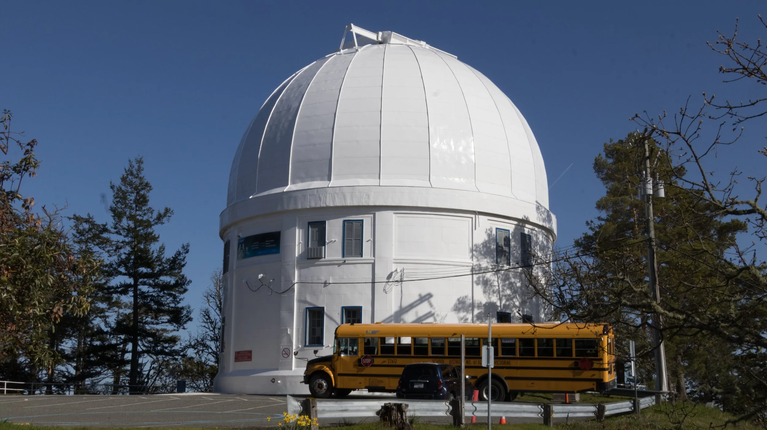 A white observatory dome with small windows, surrounded by trees and a yellow bus parked nearby, against a clear blue sky.