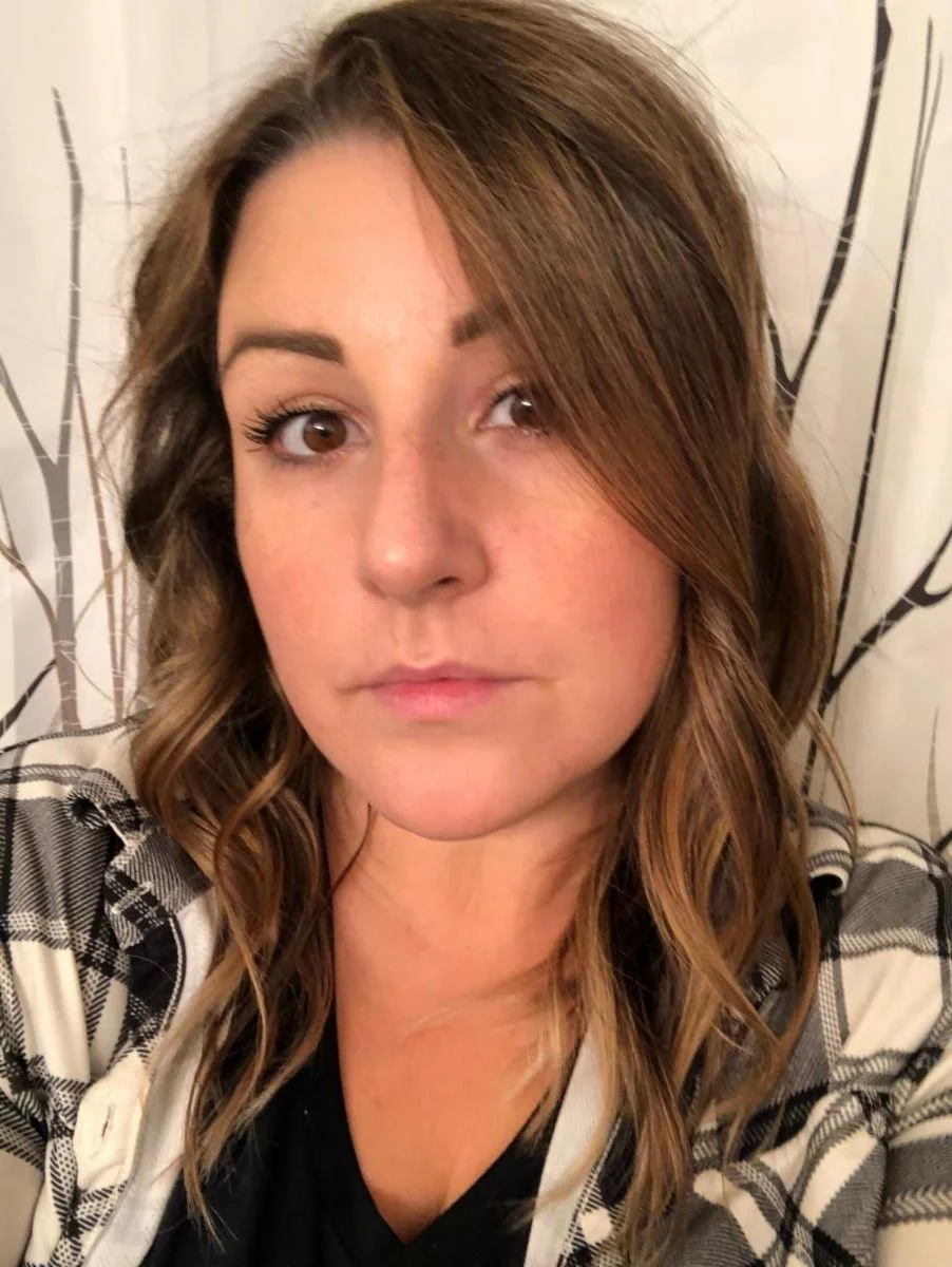Person with long brown hair wearing a plaid shirt, looking at the camera with a neutral expression.