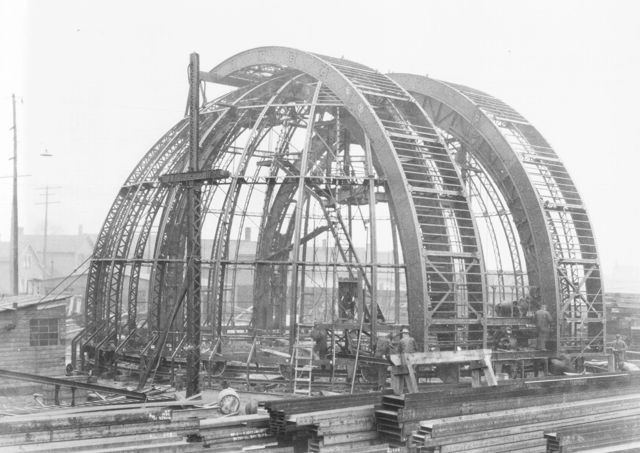 Black and white photo of a large steel structure under construction, with workers on site. The structure features multiple curved beams forming an arch-like frame. "Hale Assembling Co. No 24837" is visible on the beams.