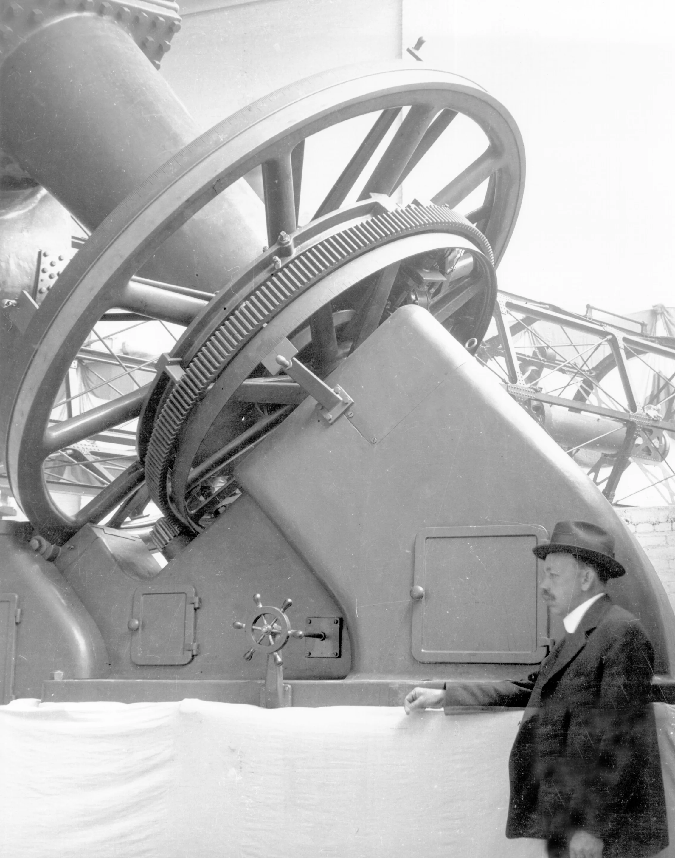 A man in a suit and hat standing next to a large industrial machine with gears and a wheel mechanism.
