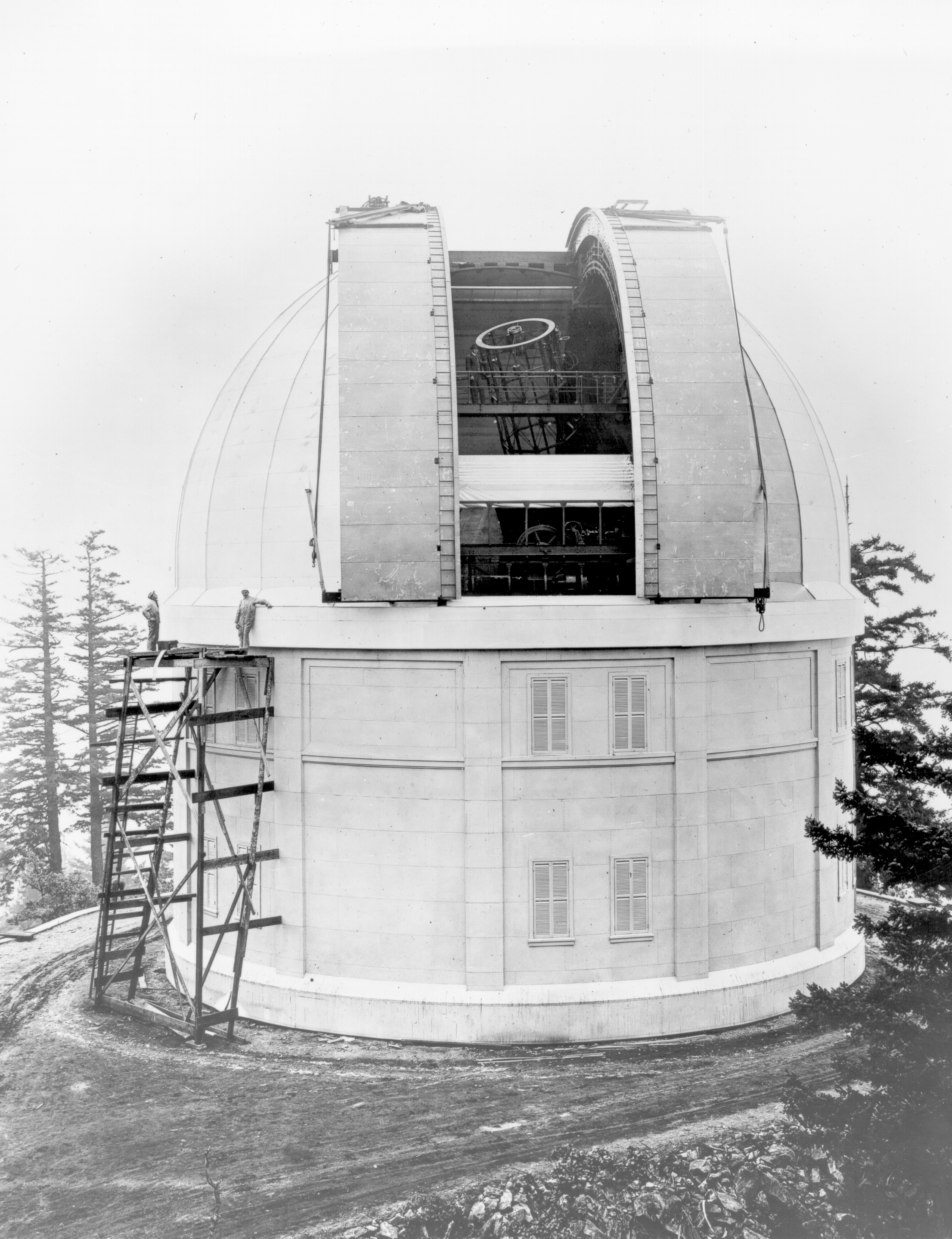 Black and white photo of a large building that looks like an observatory or a tower, with scaffolding and construction workers working on it. The structure has a curved roof, and the sky is clear. There are trees in the background.