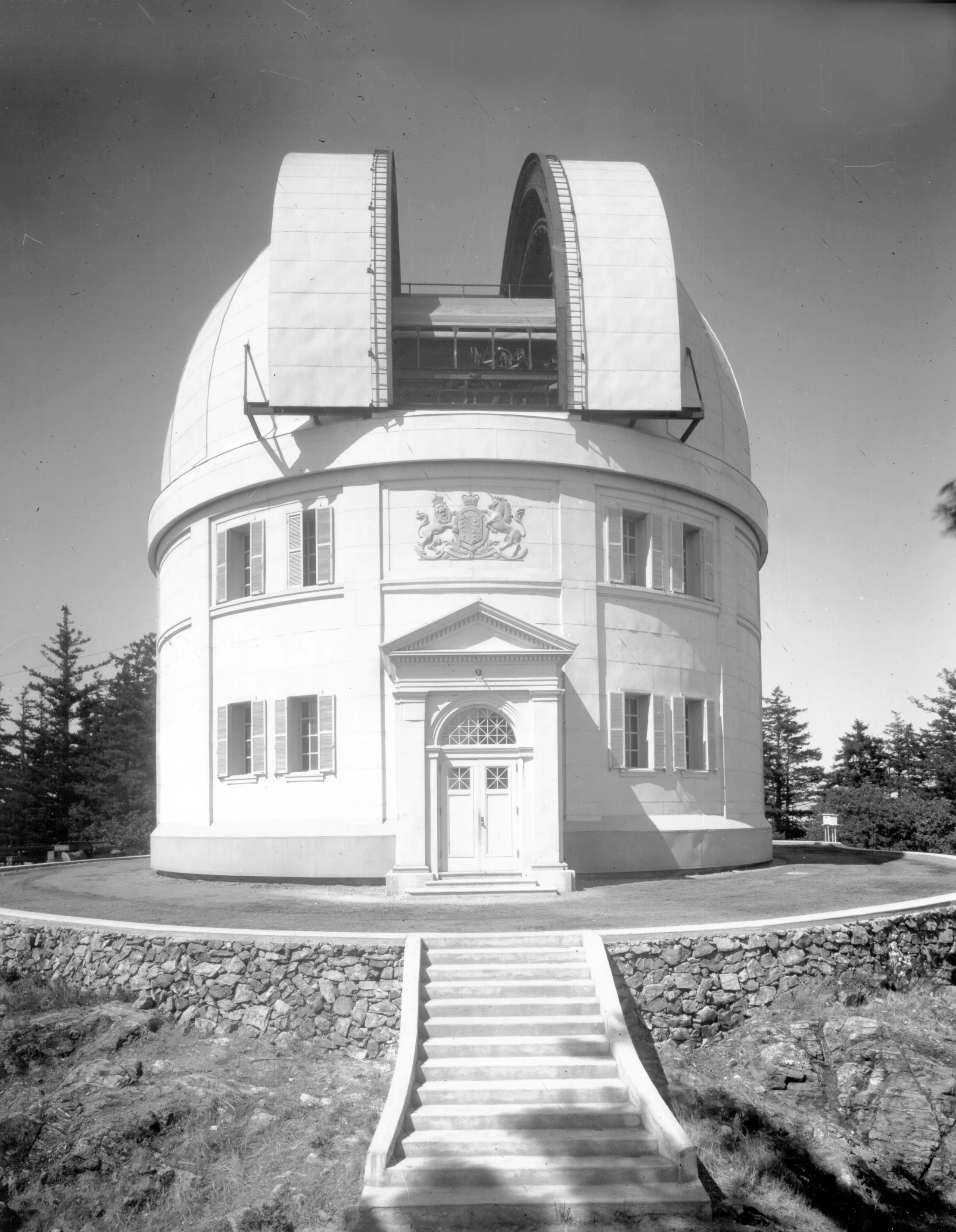Black and white photograph of a white building resembling a castle tower with a large telescope on the upper level. The building has a rounded top, decorative crests, and windows. A staircase leads up to the entrance, with trees and a partly cloudy s