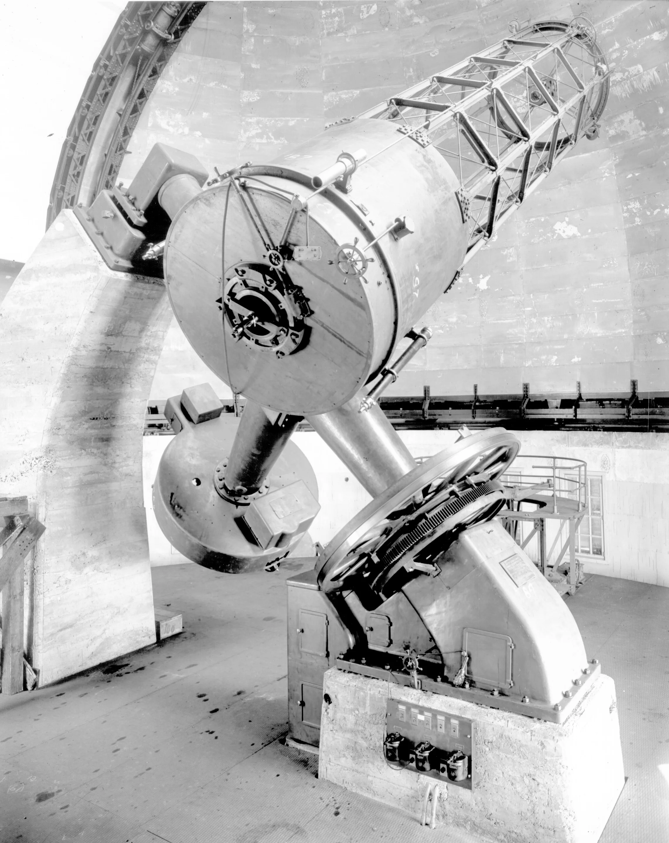 Black and white photo of a large telescope inside an observatory with a curved ceiling, several control panels, and support structures.